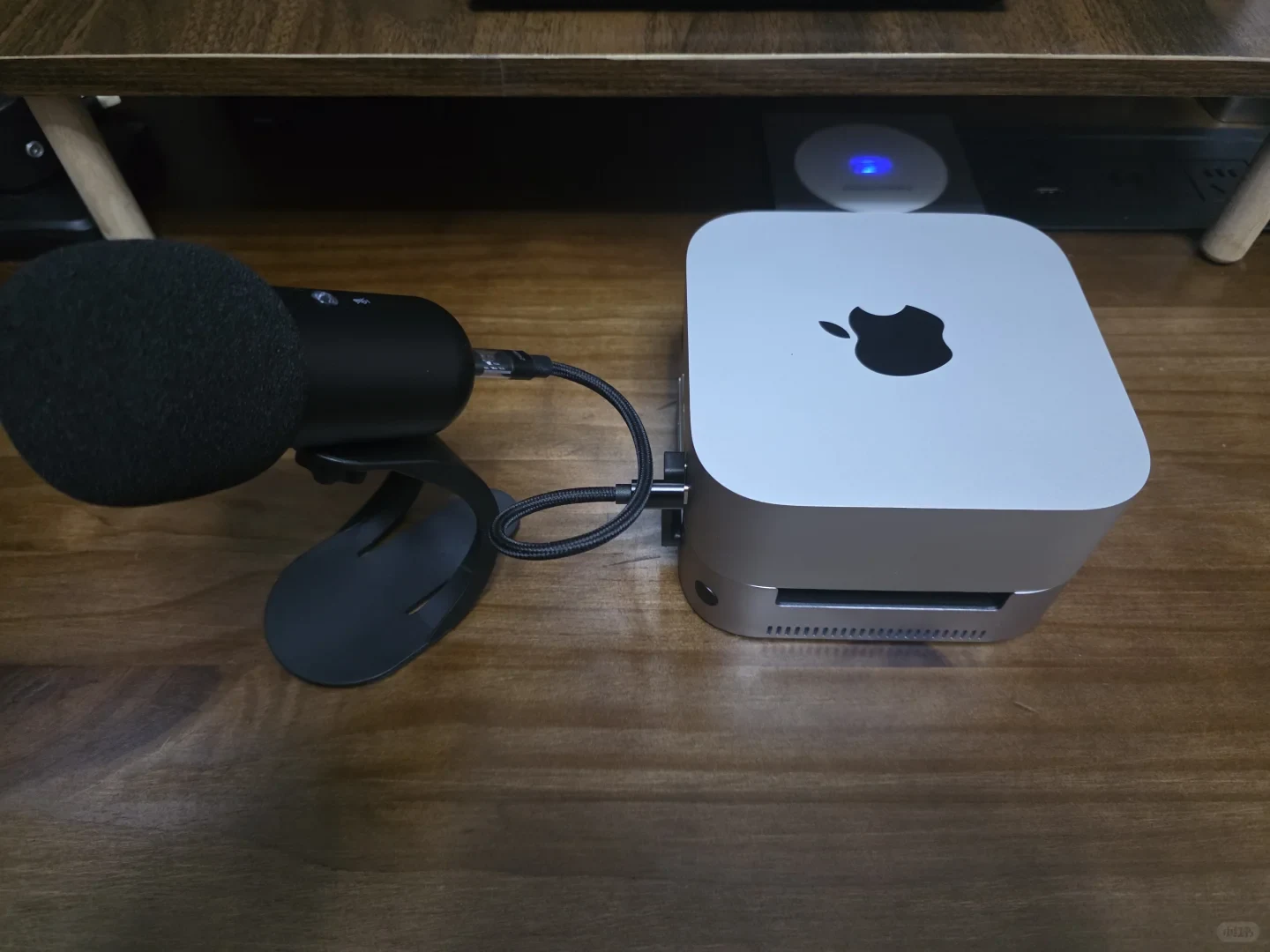 A silver Apple Mac mini sits on a wooden desk beside a black microphone on a stand, connected by a short braided cable. A small device with a blue light is visible behind them.