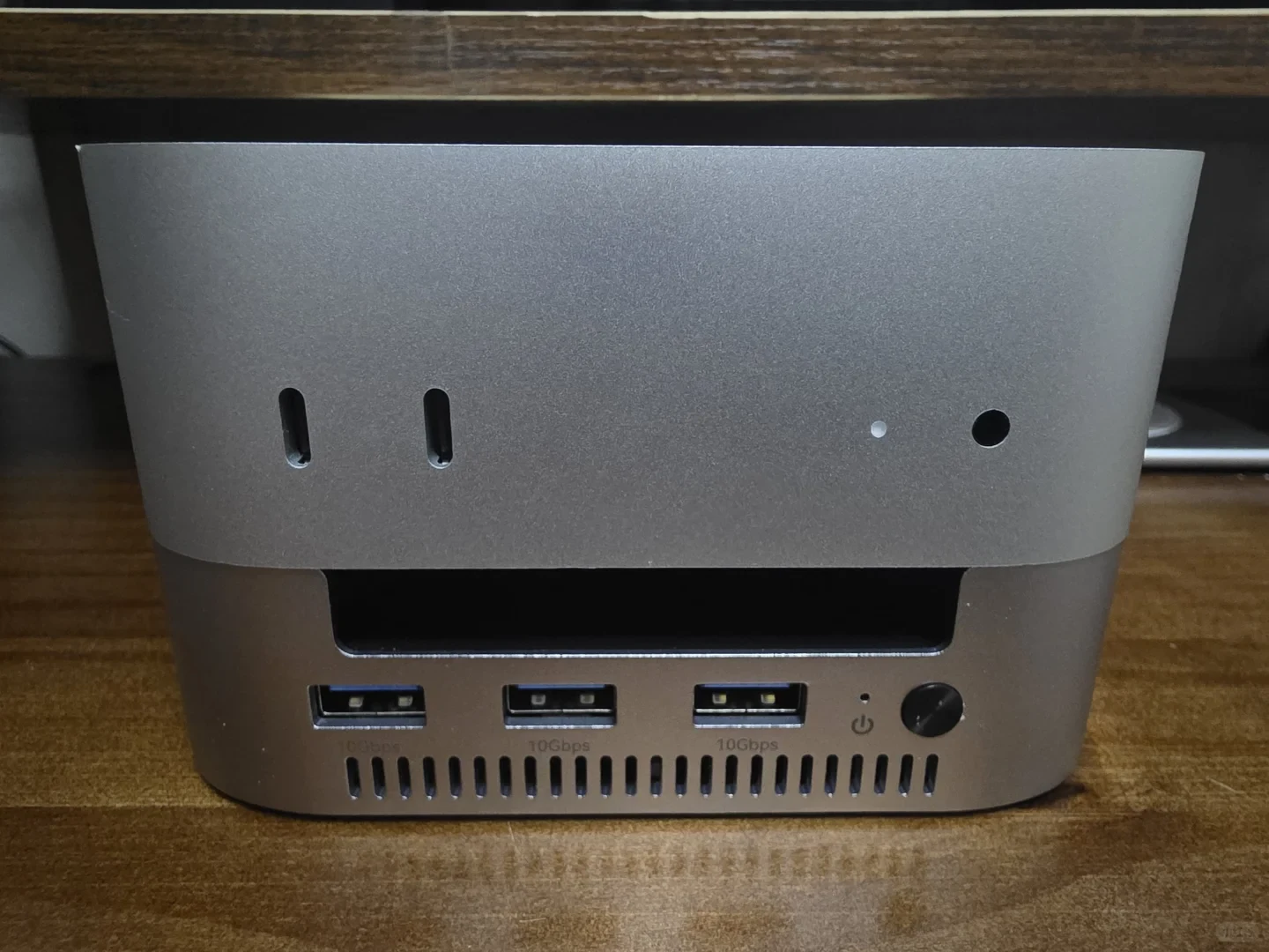 A compact silver desktop computer sits on a wooden surface, viewed from the front. It has two vertical USB-C ports, two small circular openings, a wide slot, three USB-A ports, ventilation slits, and a power button; faint “10Gbps” labels appear below two ports.