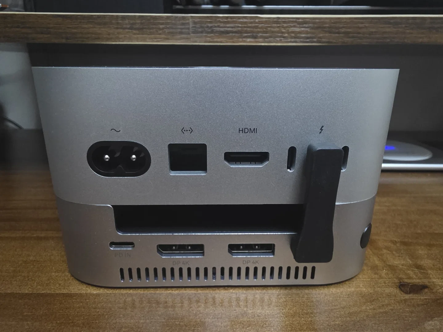 Rear view of a Mac mini on the ACASIS dock on a wooden desk. A short black host connector links the computer and dock; two DisplayPort sockets, a PD input and ventilation slots are visible on the dock.