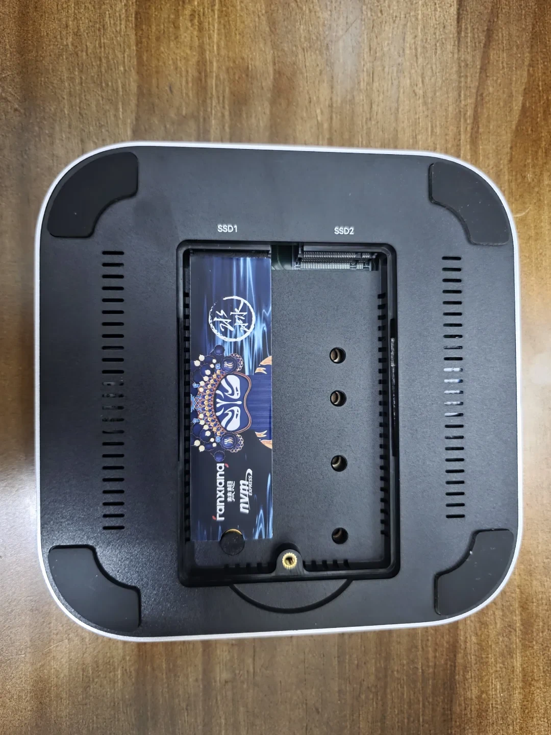 Underside of a black, square device on a wooden surface, with its cover removed to show two SSD slots. One slot contains a blue-labeled SSD; the other is empty. The slots are labeled “SSD1” and “SSD2.”