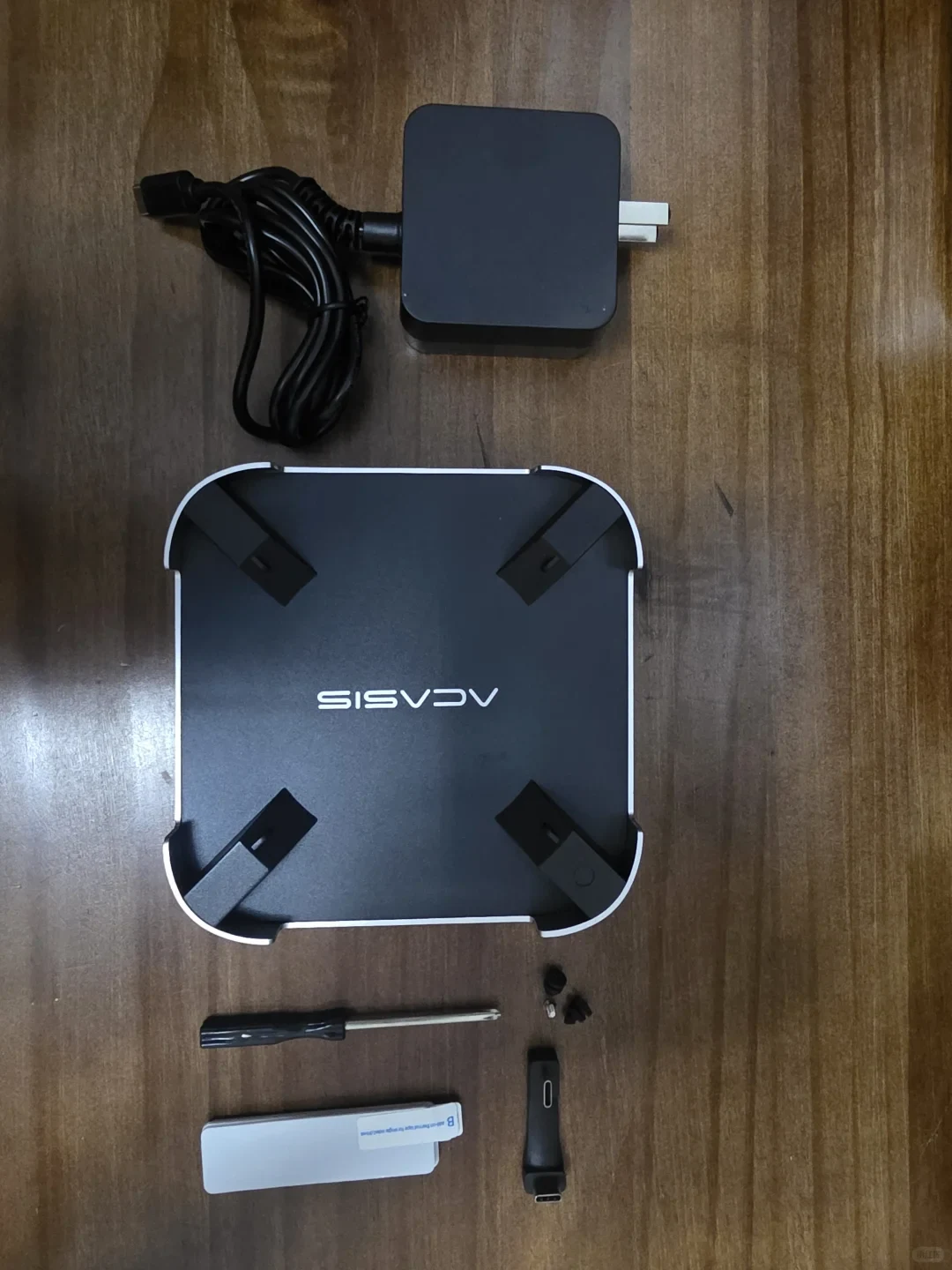 A square black device with four corner clips and white trim lies on a wooden surface, surrounded by a power adapter and coiled cable, a screwdriver, small screws, a slim black adapter, and a folded white card. The device bears a stylized logo.