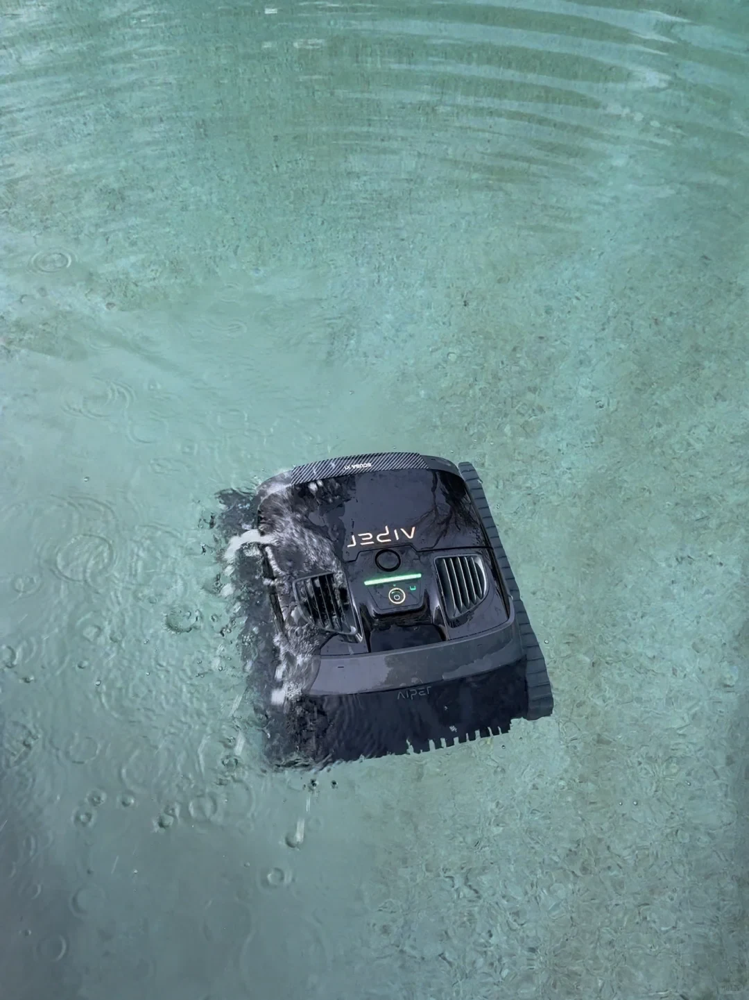 A black Aiper robotic pool cleaner submerged in clear swimming pool water with water ripples on the surface.