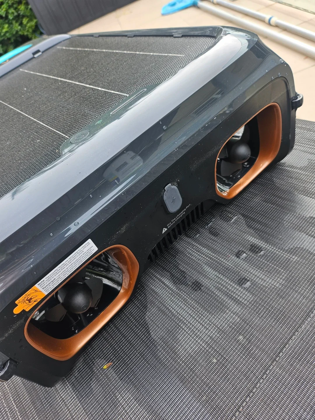 Close-up of a dark grey solar-powered pool skimmer robot resting outdoors on a mesh surface, showing two copper-rimmed propeller tunnels, a central rubber-covered charging port, and a top solar panel surface.