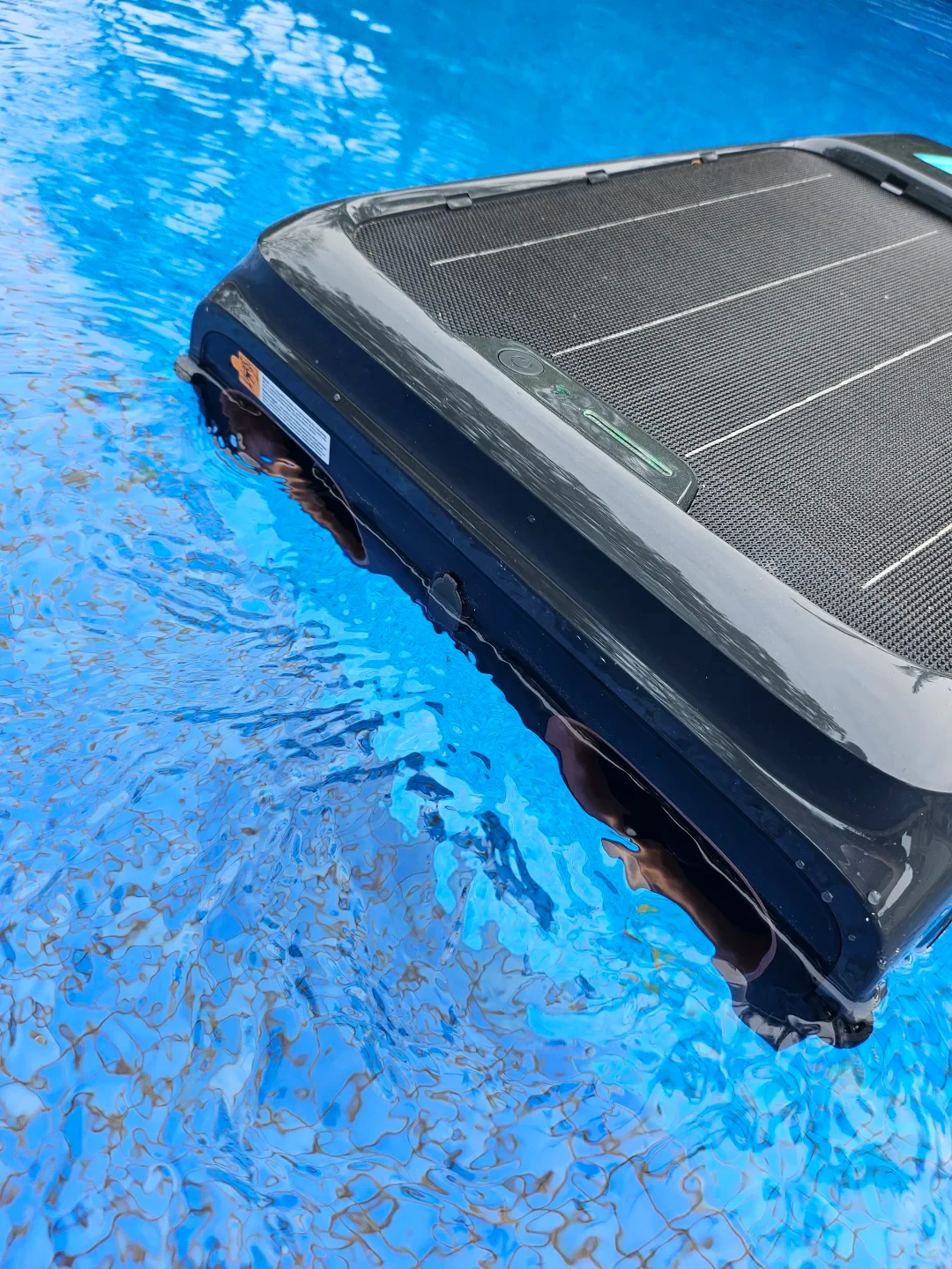 A black solar-powered robotic pool skimmer floating on clear blue rippling swimming pool water.