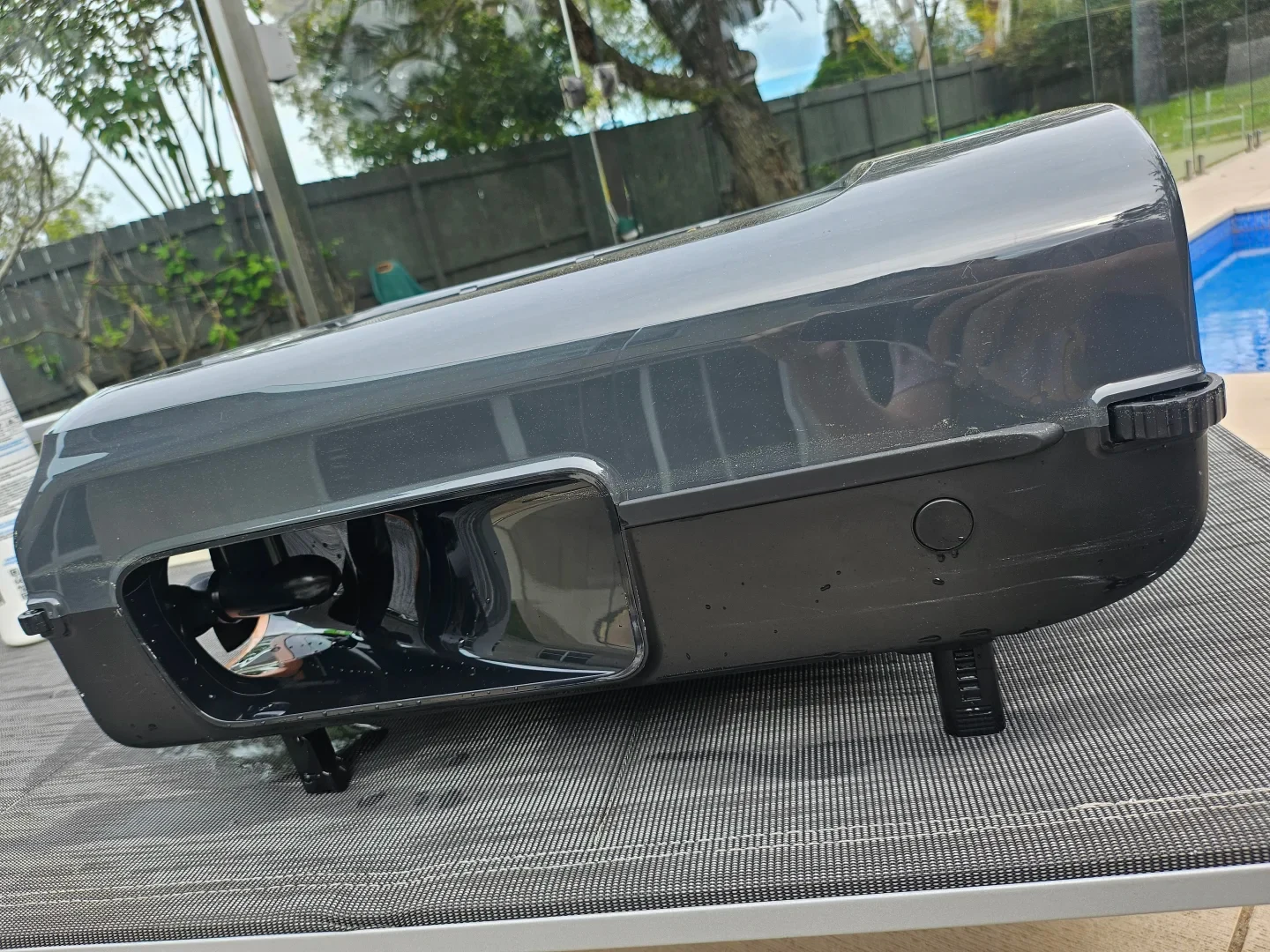 A side view of a dark grey and black robotic pool cleaner resting on an outdoor mesh table next to a swimming pool, showing an internal propeller tunnel and supporting feet.