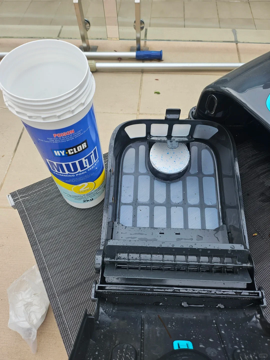 An open container of Hy-Clor Multi swimming pool tablets stands next to the opened black filter basket of a robotic pool cleaner, which holds a speckled white tablet in its top compartment.