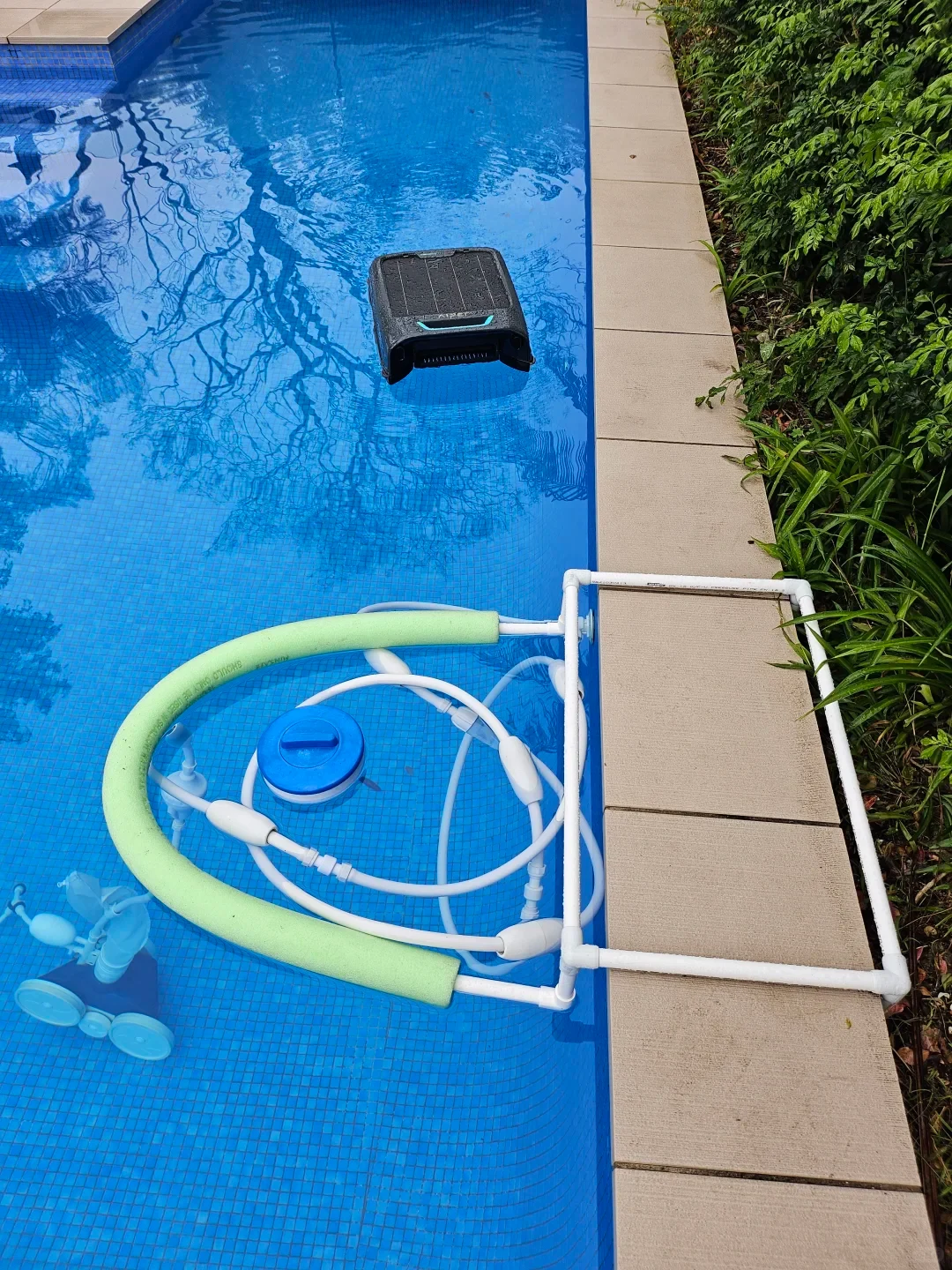 A high-angle view of a backyard swimming pool with blue tiled water. A black robotic surface skimmer floats on the water toward the top, while in the foreground, a white PVC pipe frame fitted with a curved light green pool noodle floats along the edge, tethering white pool cleaner hoses and a blue floating dispenser.