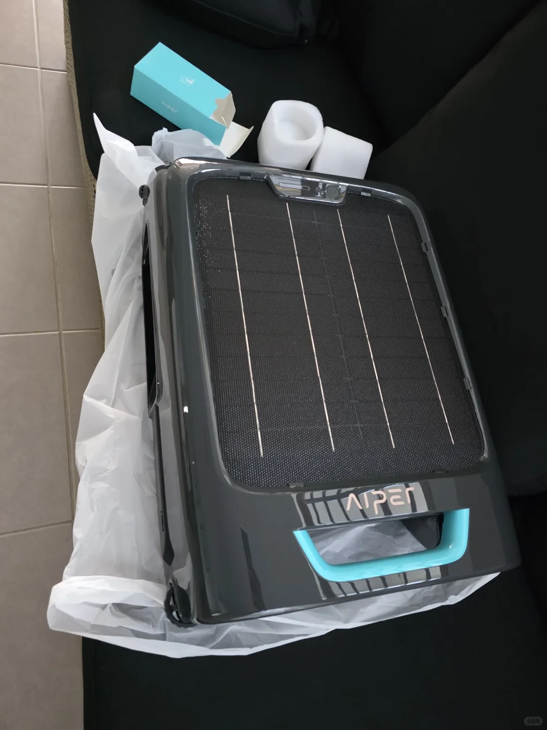 A top-down view of a dark gray Aiper solar-powered robotic pool skimmer unboxed onto white protective plastic wrap. The top surface displays a solar panel with vertical silver contact lines, accompanied by a light blue carrying handle and the Aiper logo at the base. A small teal accessory box and white foam packing cushions rest in the background on a black surface.