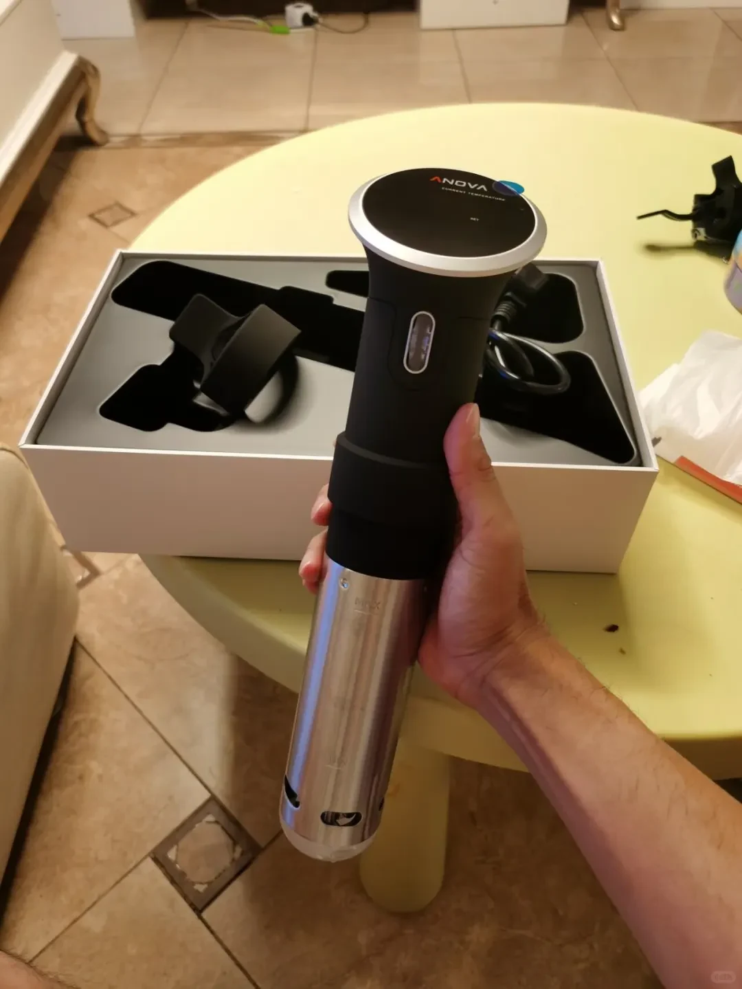 A person holds an Anova sous vide immersion circulator with a black upper body and stainless steel heating shaft over its open white packaging box.