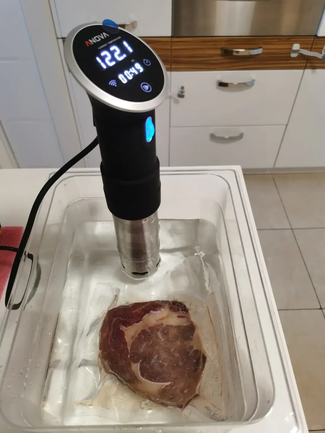 An Anova precision immersion circulator mounted in a clear plastic water bath container, cooking a vacuum-sealed steak immersed at the bottom.