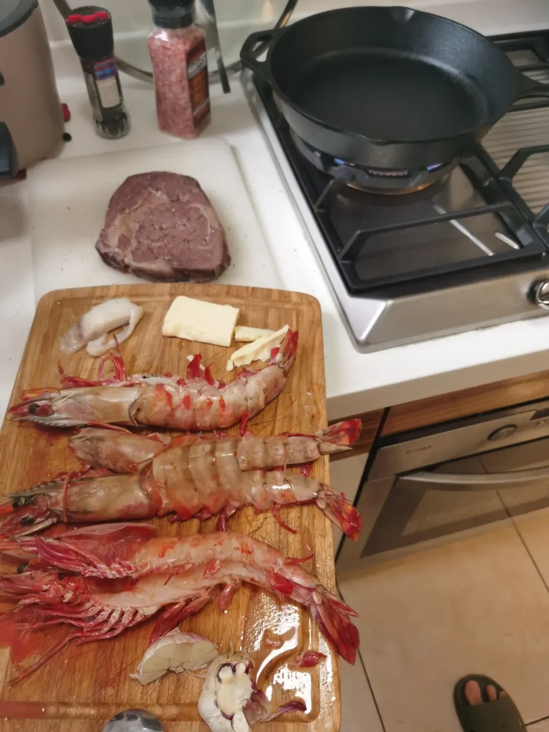 A kitchen counter setting with a wooden cutting board holding large split prawns, butter, and garlic, next to a sous-vide cooked thick steak and a black cast iron skillet heating over a gas stove flame.