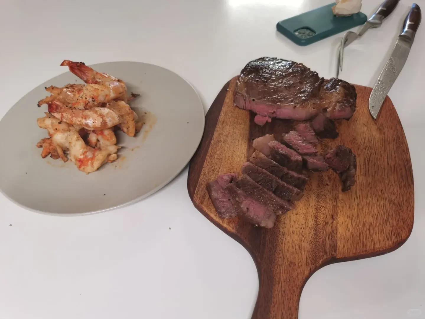 A sliced, seared thick-cut steak showing a rare pink interior on a wooden cutting board next to a plate of cooked shrimp, a carving knife, and a fork.