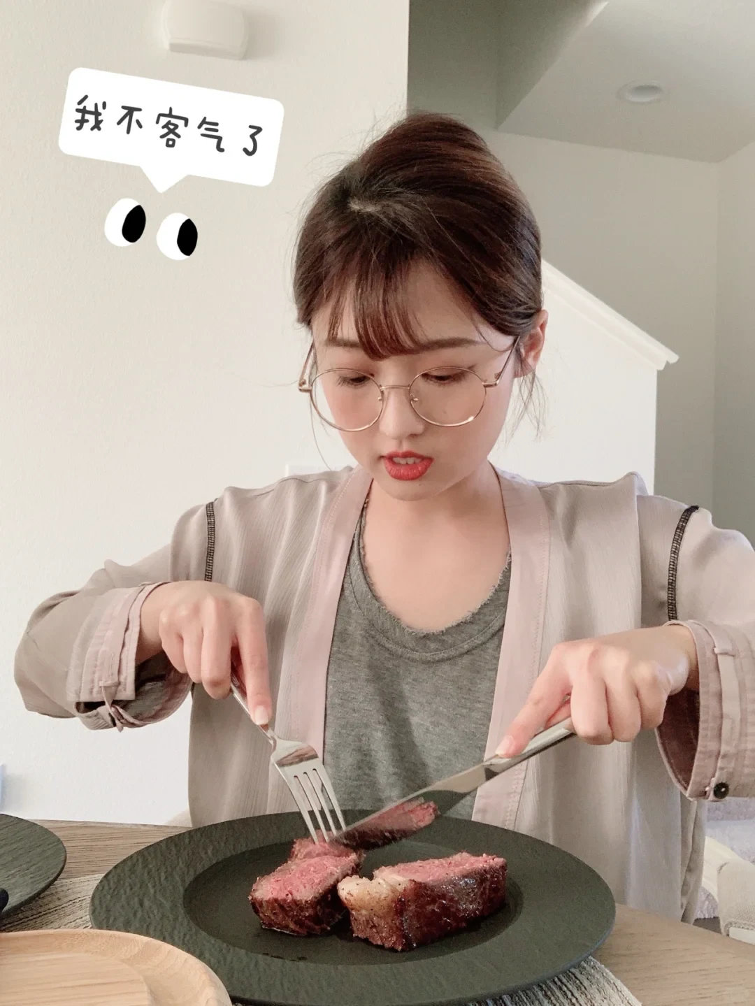 A young woman with round glasses cuts into a thick slice of medium-rare steak on a black plate using a fork and knife. A speech bubble sticker in the top-left corner contains Chinese text next to a cartoon eyes sticker.