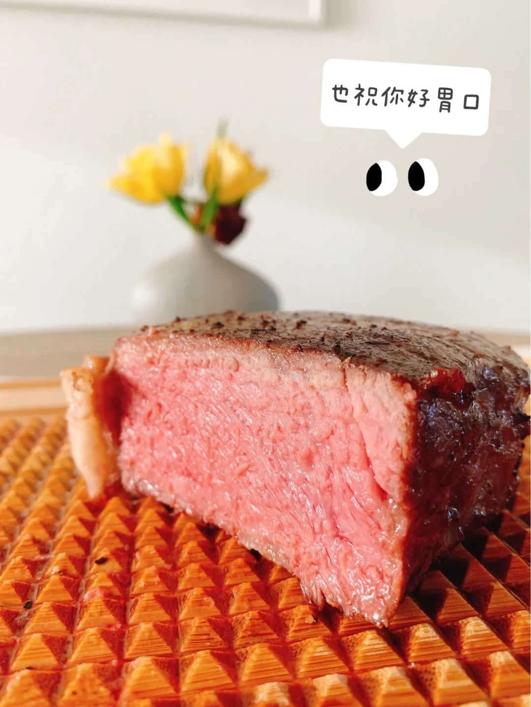 A close-up shot of a thick cut of medium-rare steak with a seared crust resting on a textured wooden cutting board, with a vase of yellow flowers softly blurred in the background and a speech bubble graphic above the steak.