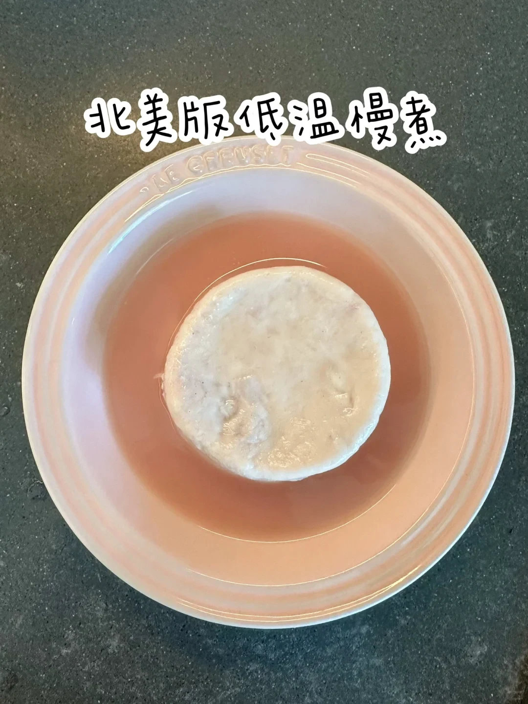 A round, pale cooked meat patty resting in light pink broth inside a pink ceramic Le Creuset pet bowl on a dark grey countertop, with Chinese text overlay at the top.