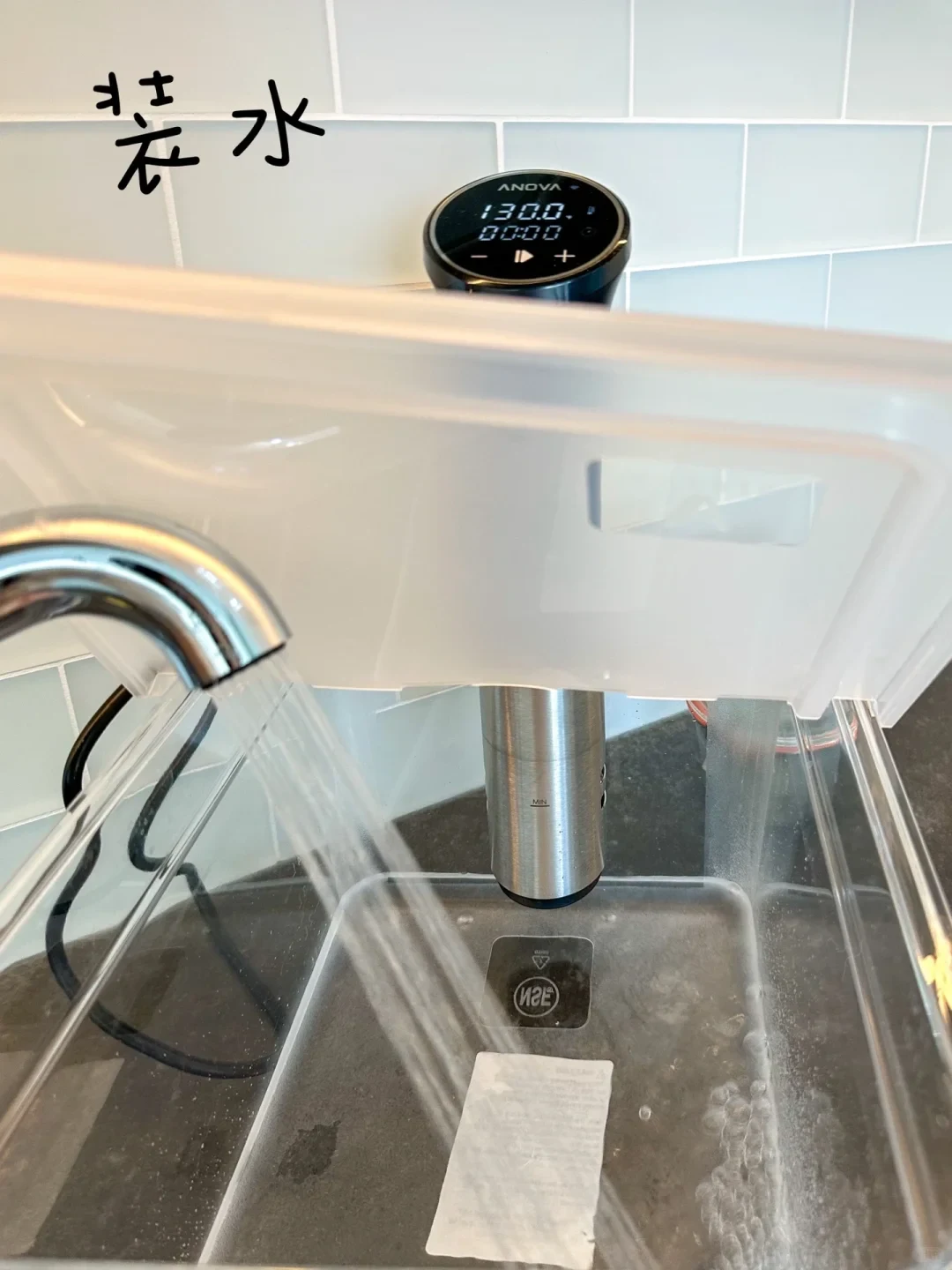 A clear plastic sous vide container with an Anova precision cooker mounted inside is being filled with tap water from a kitchen faucet.