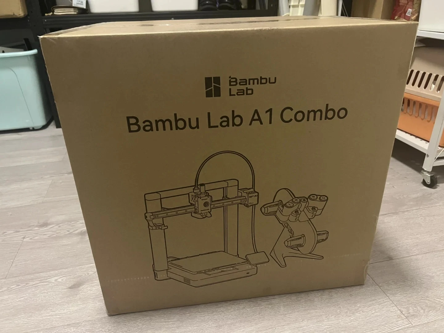 A large brown cardboard shipping box printed with the Bambu Lab logo, 'Bambu Lab A1 Combo', and line drawings of the A1 3D printer and AMS lite accessory, sitting on a light-colored wood floor.