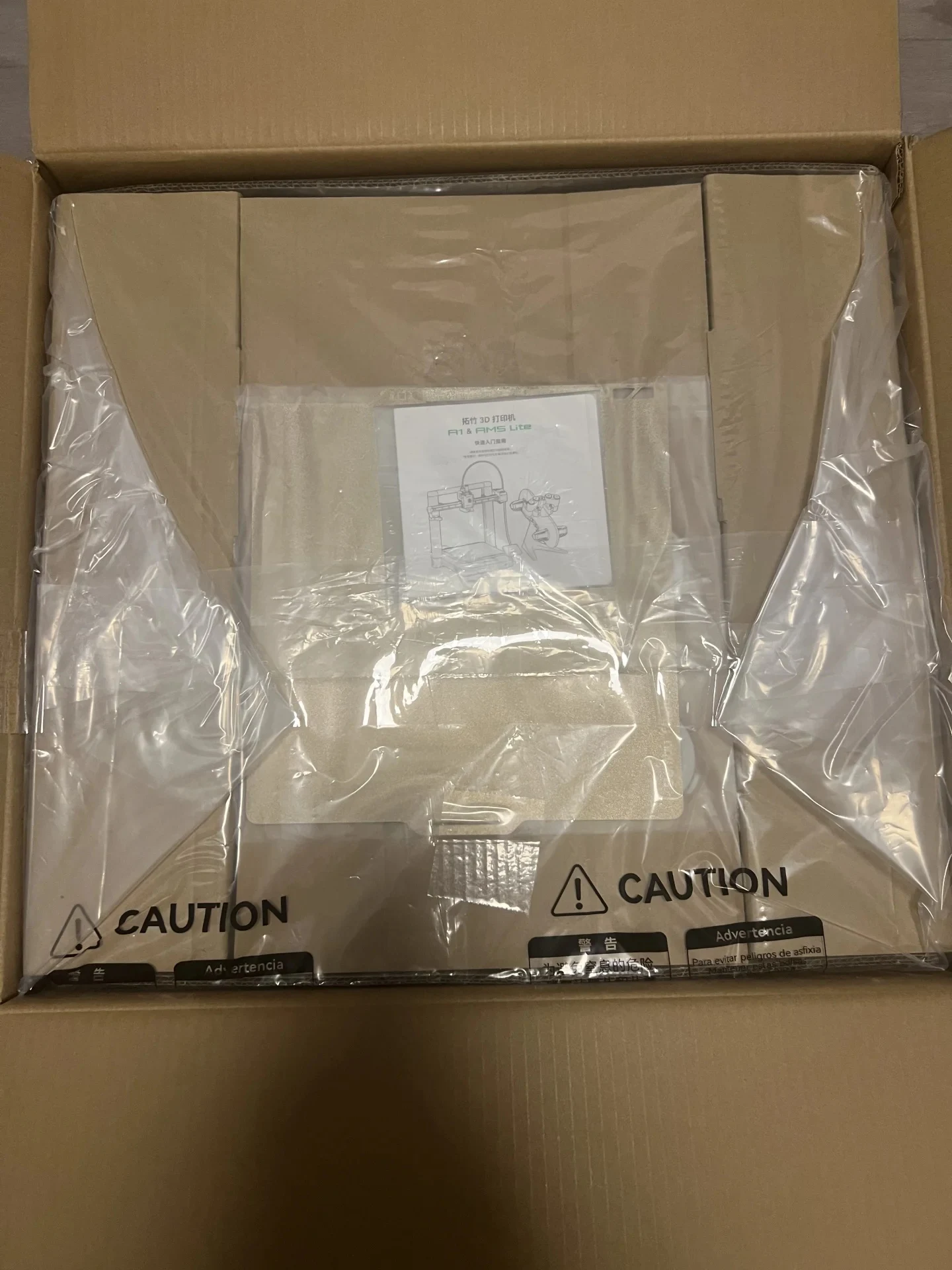 Top-down view into an opened cardboard shipping box containing the Bambu Lab A1 and AMS Lite packaging sealed beneath clear plastic wrap, with a quick start guide placed in the center.