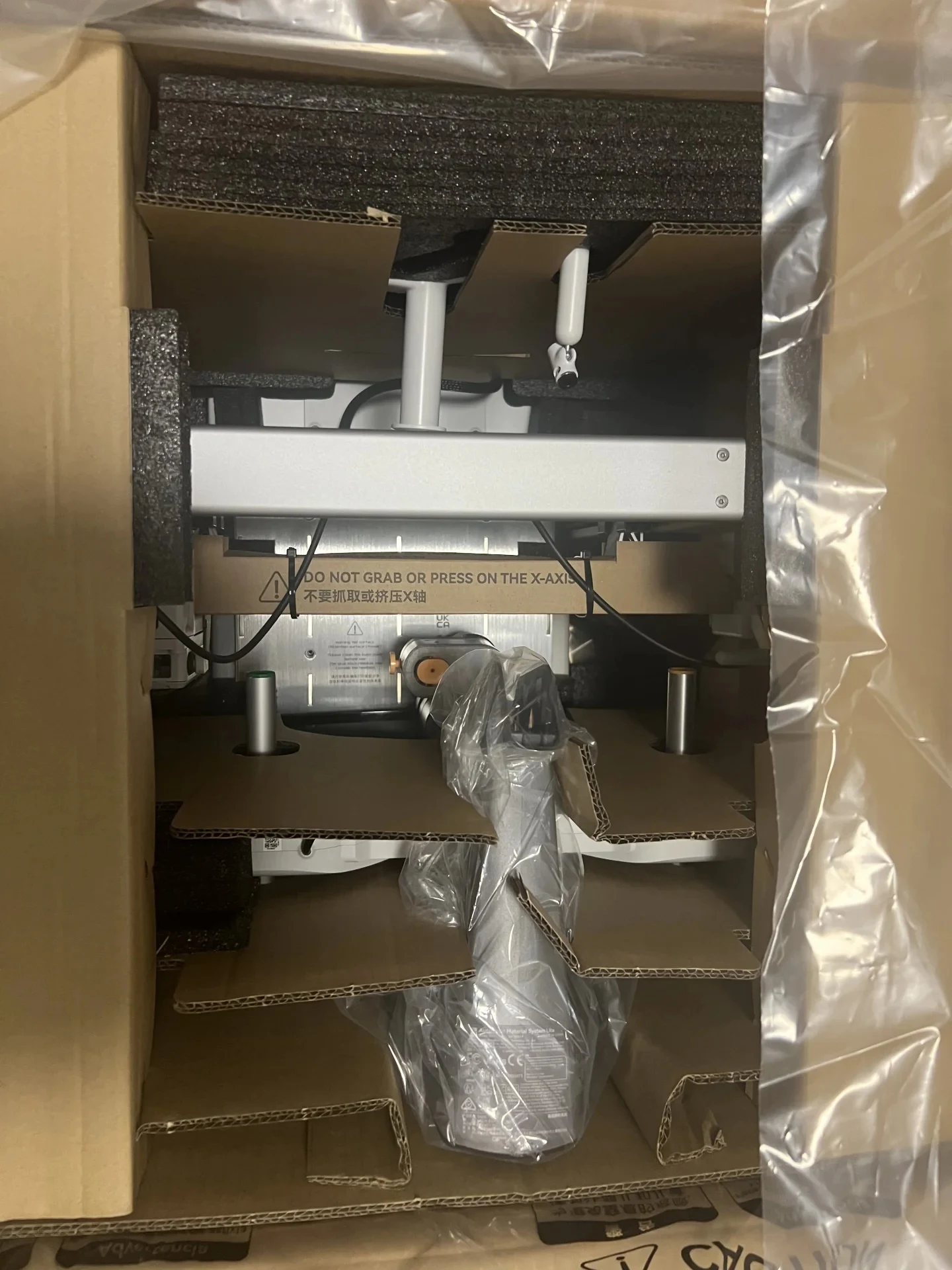 Top-down view into an open cardboard shipping box containing a Bambu Lab A1 3D printer secured by molded cardboard inserts and dark foam padding, with a warning strip spanning the gantry.