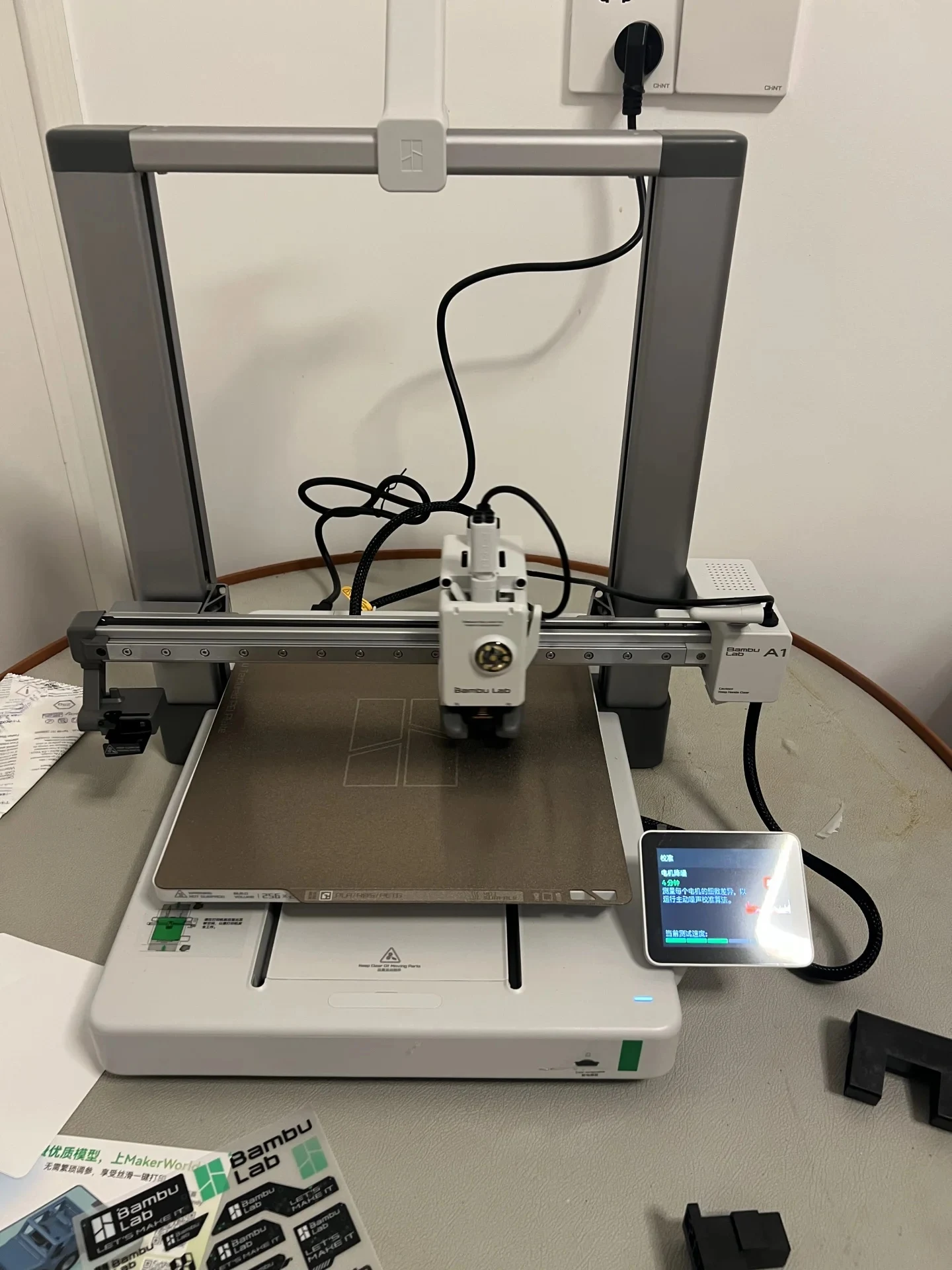 A Bambu Lab A1 3D printer set up on a table, with its printhead centered above the textured PEI build plate and the small touchscreen displaying a motor noise cancellation calibration sequence.