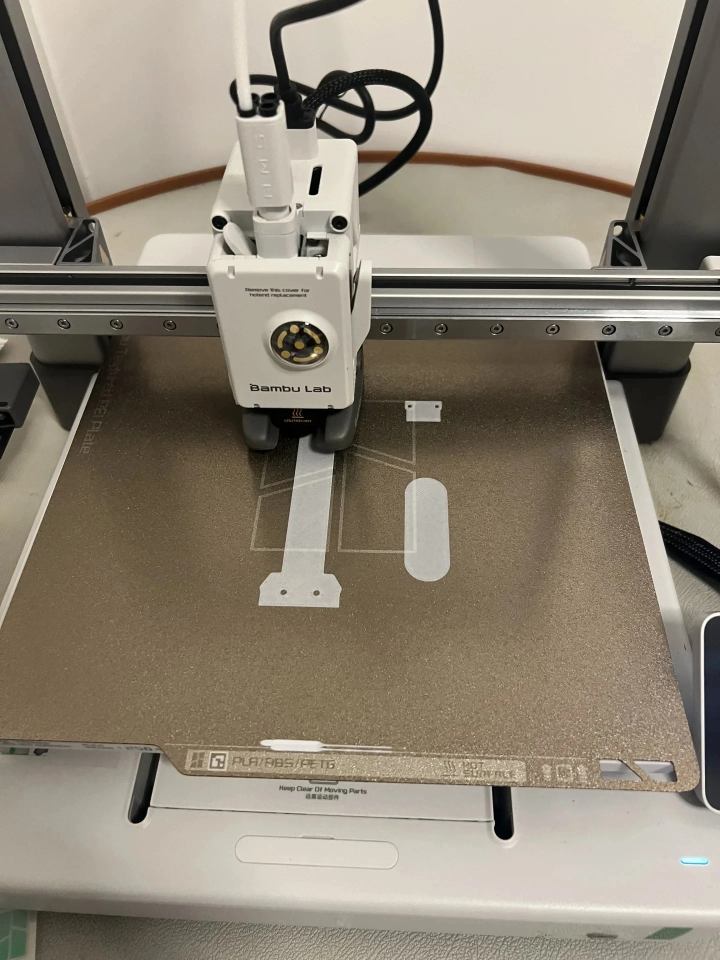 Top-down view of a Bambu Lab A1 3D printer toolhead depositing white filament onto a textured PEI build plate.