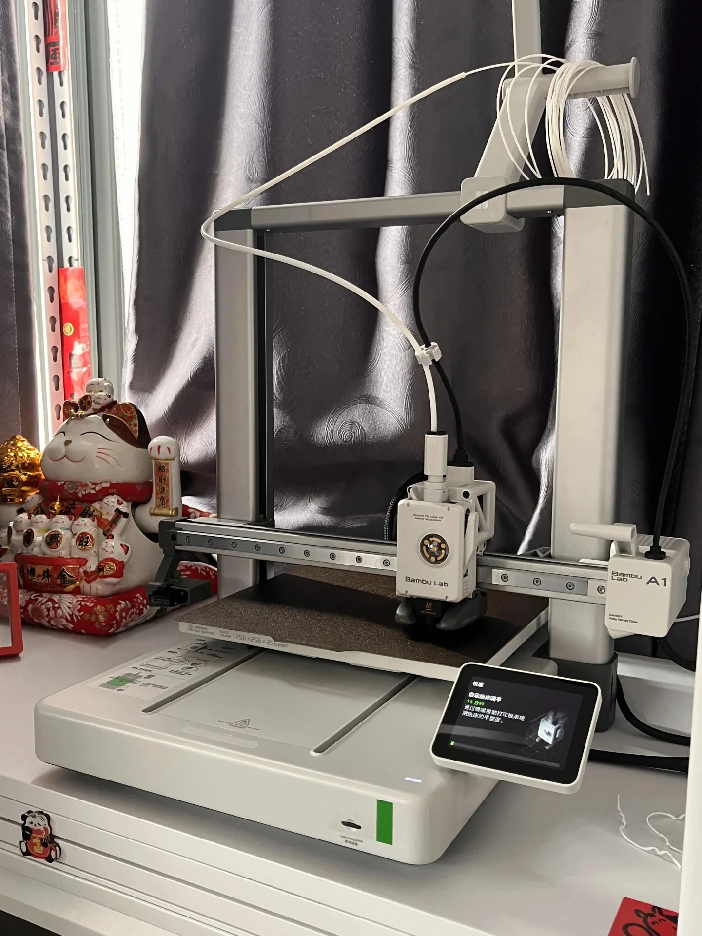 A Bambu Lab A1 3D printer assembled on a white shelf next to a lucky cat figurine, with its touchscreen displaying an automatic bed leveling calibration in progress.