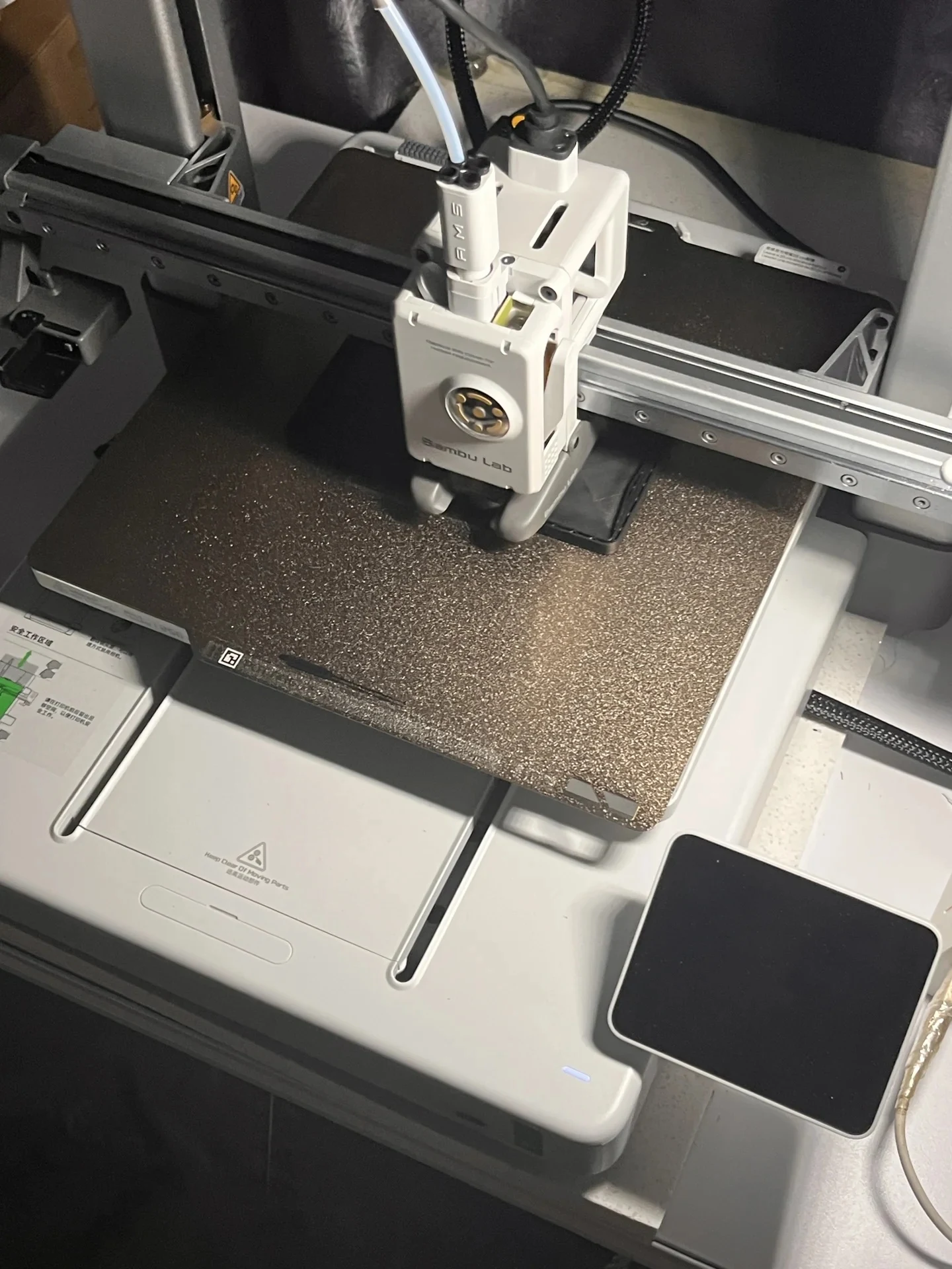 Top-down view of a Bambu Lab A1 3D printer with its toolhead actively printing a black plastic object on the textured build plate.