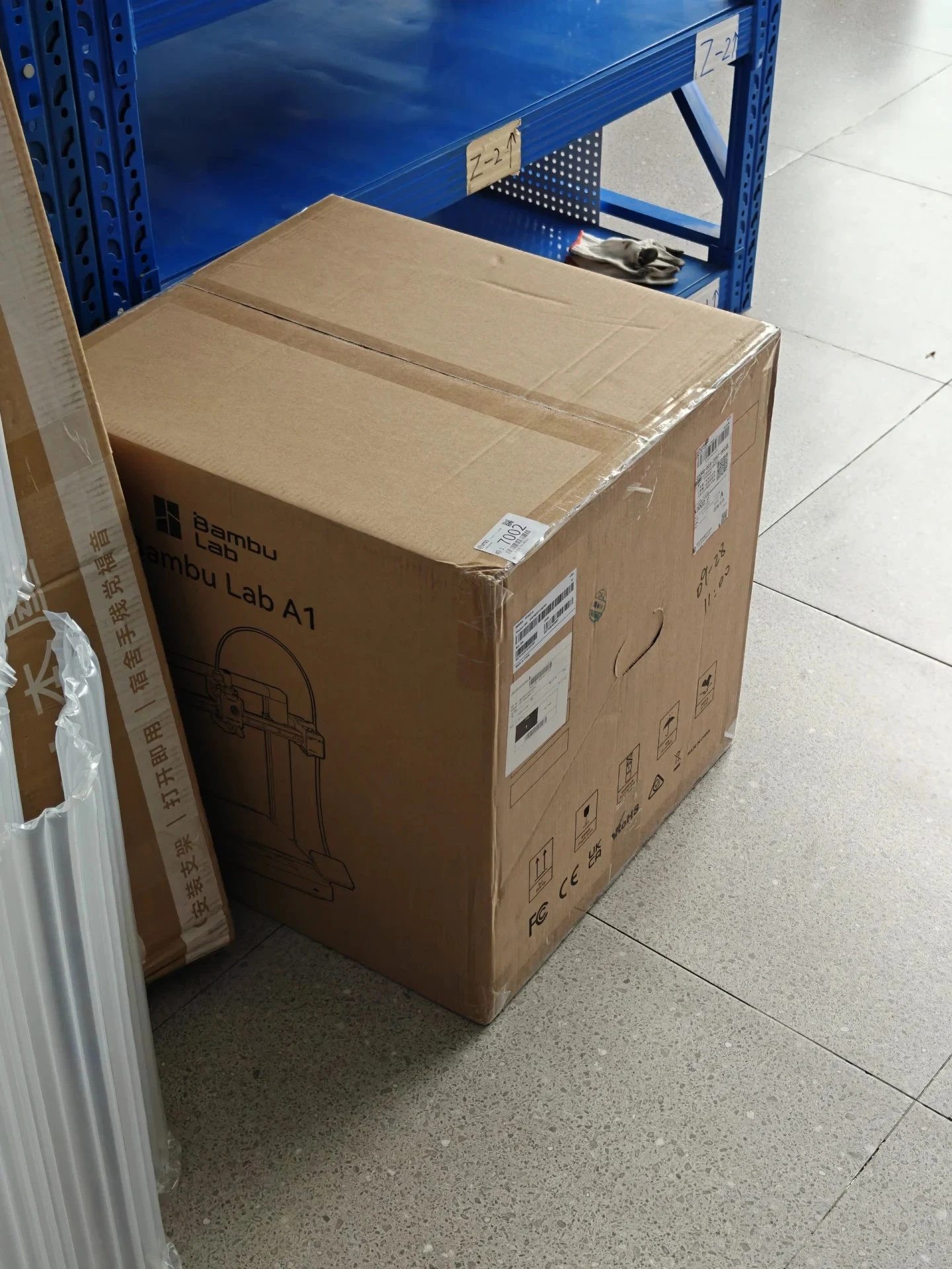 A large sealed cardboard box for a Bambu Lab A1 3D printer rests on a tiled floor beside blue shelving and packaging materials.