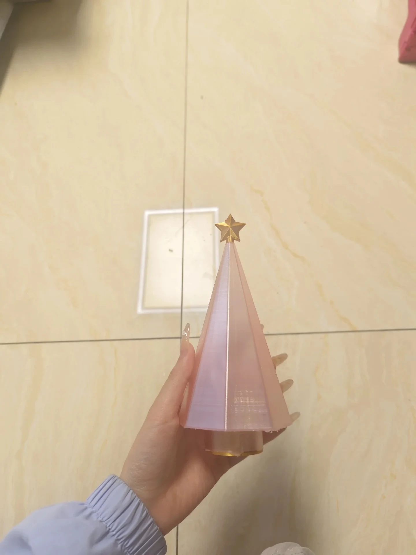 A hand holding a 3D-printed geometric Christmas tree figurine with faceted translucent pink panels and a gold star on top, held above a tiled floor.
