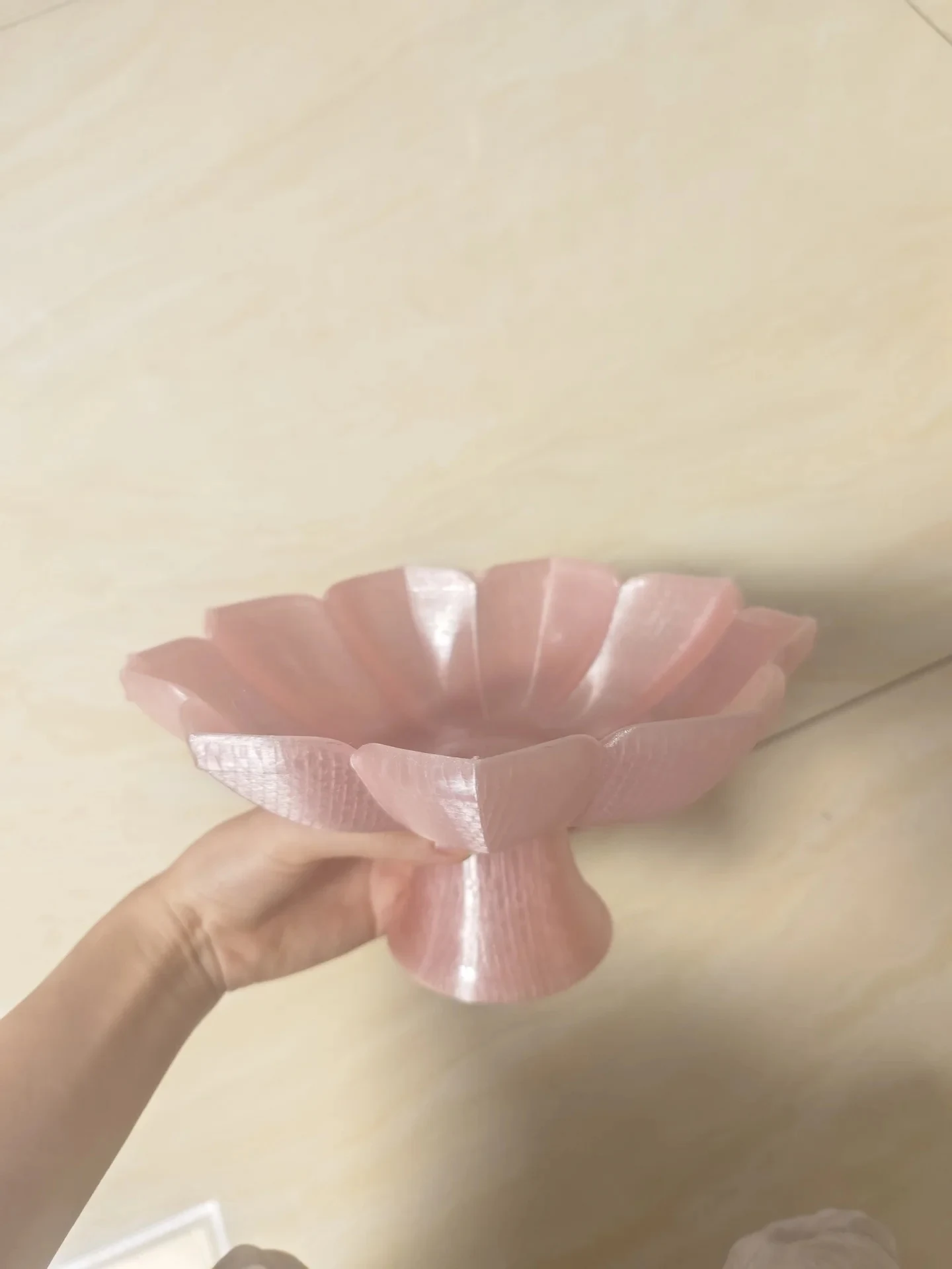 A hand holds a 3D-printed lotus petal pedestal dish crafted from shimmering, translucent pink filament, displaying fine layer lines across its flared petals and base.