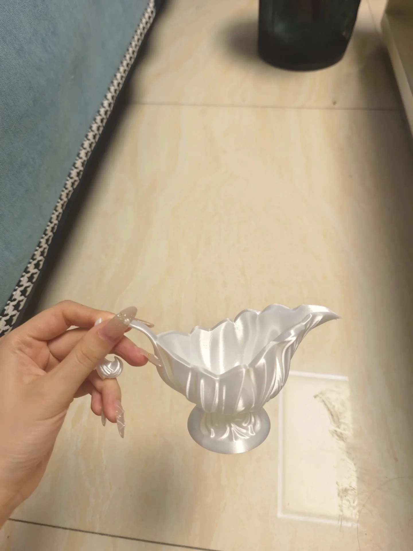 A hand holds an ornate, 3D-printed white footed bowl with a flared spout and a decorative handle over a light-colored tile floor.