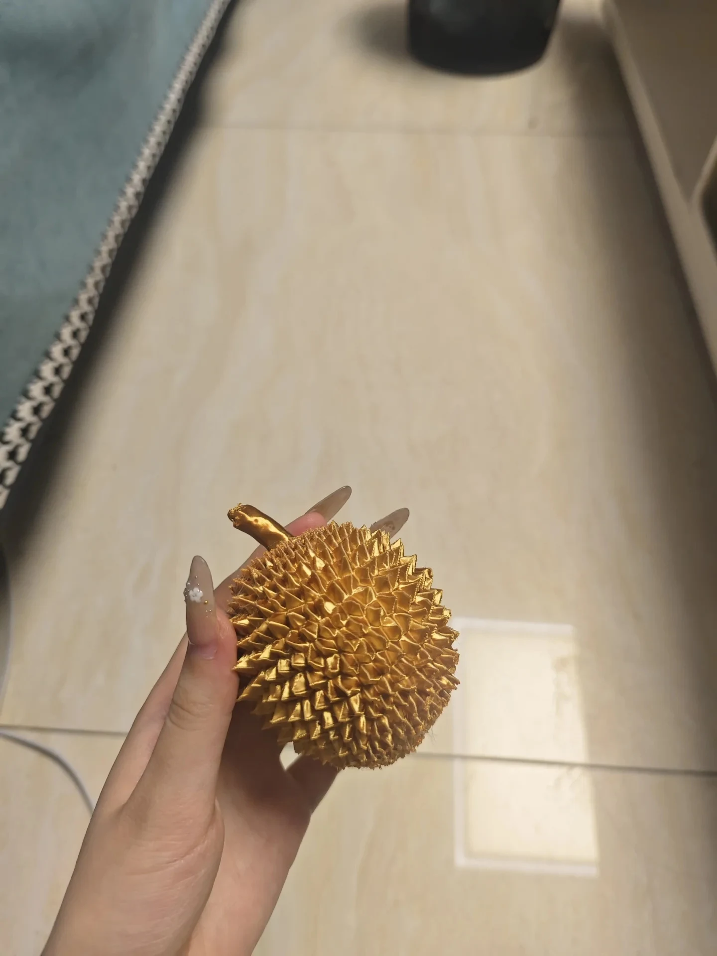 A hand holding a small gold-colored 3D-printed spiky durian model over a tiled floor.