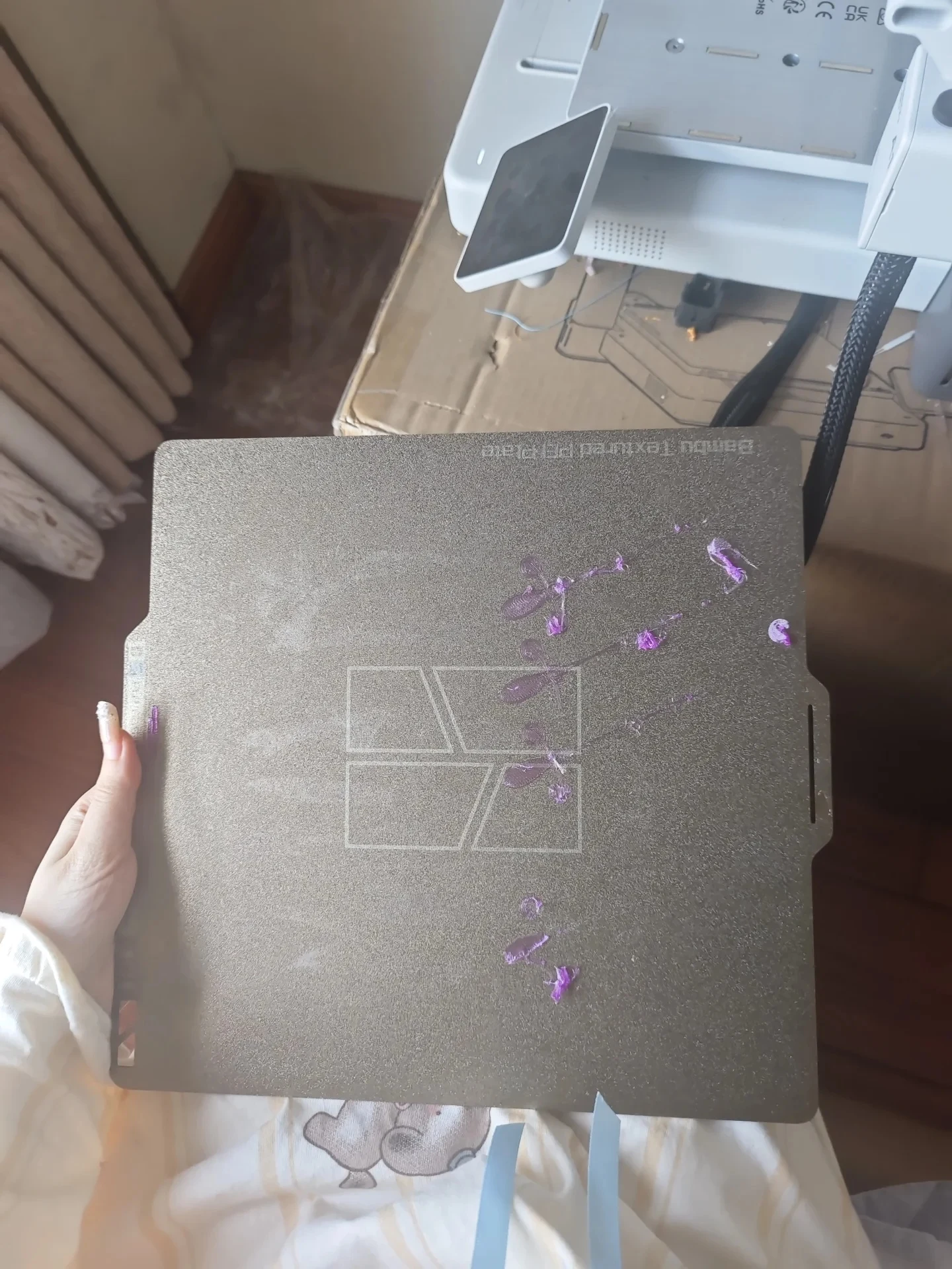 A hand holding a Bambu textured PEI build plate marked with purple filament residue, with the base and touchscreen of a 3D printer in the background.