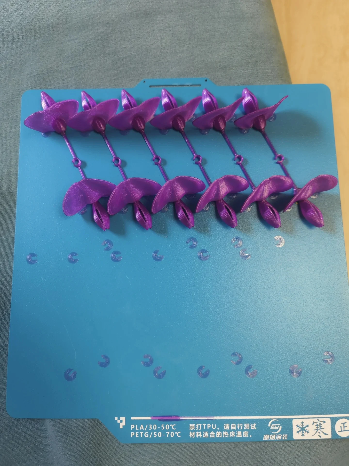 Top-down view of a light blue 3D printer build plate holding rows of glossy purple 3D-printed flower petal parts and printed circular skirt remnants, with temperature specifications labeled on the lower margin.