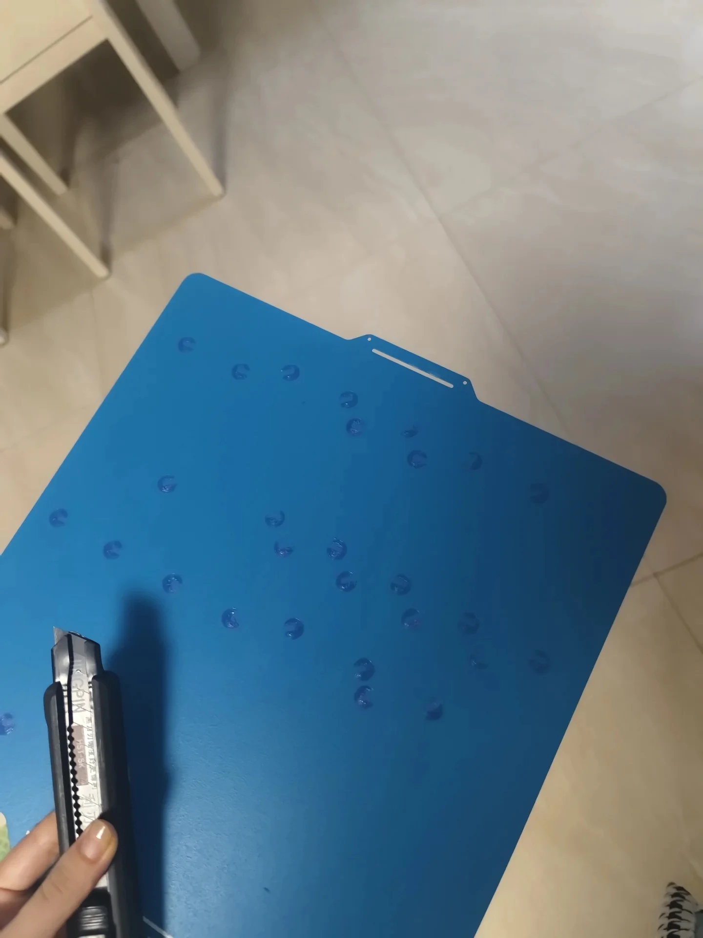 A hand holding a blue flexible 3D printer build plate covered with tiny stuck circular prints, with a utility knife held alongside it to pry them loose.