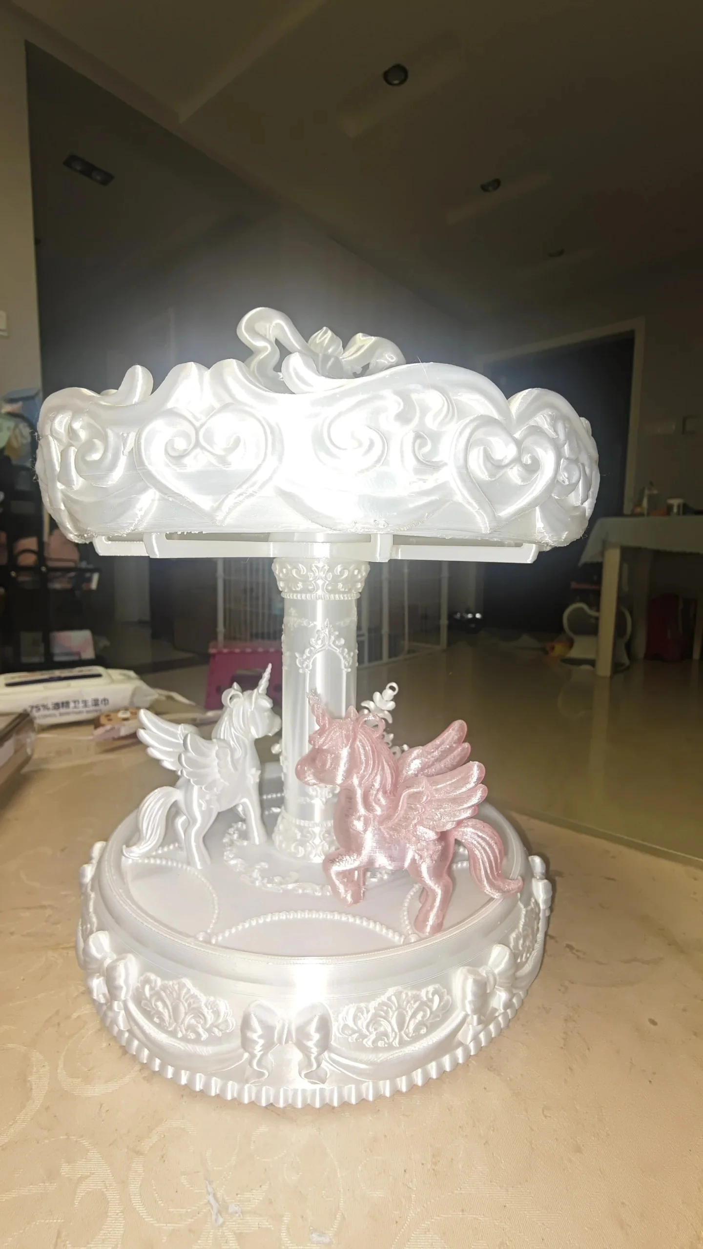 A 3D-printed ornate miniature carousel made of shiny white filament, featuring a central column supporting a decorative canopy over two winged unicorn figures, one white and one shimmering pink.