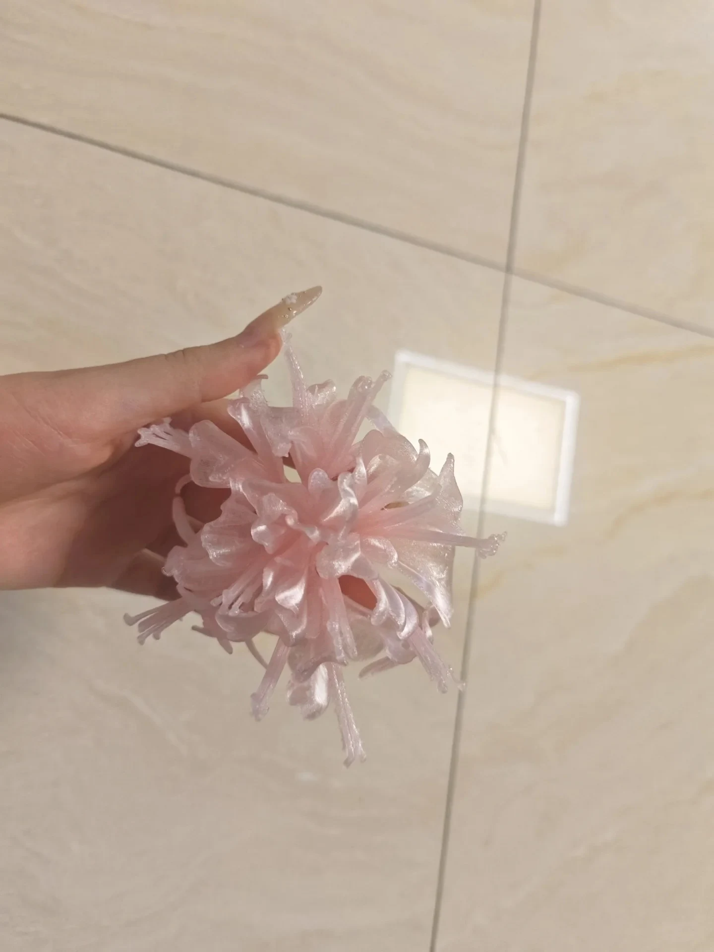 A hand holding a 3D-printed flower ornament with intricate, translucent pink petals against a neutral tiled background.