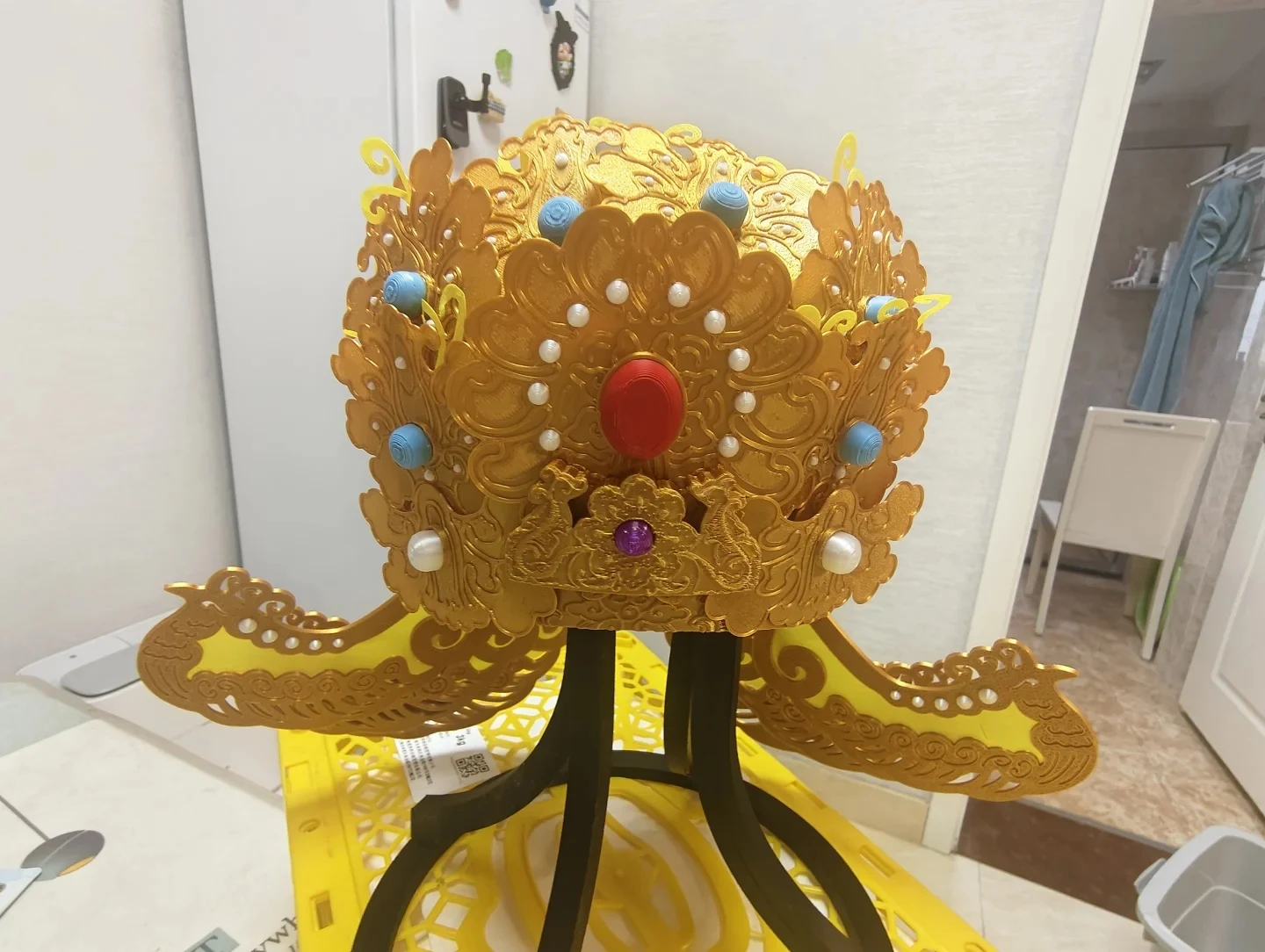 A multi-colored, ornate 3D-printed traditional Chinese crown mounted on a black stand resting on a yellow plastic crate. The golden headdress features intricate filigree patterns adorned with printed red, purple, turquoise, and pearl-like studs.