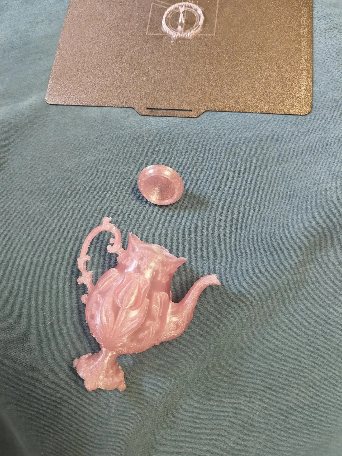 A small 3D-printed pink decorative teapot and separate lid lying on fabric next to a textured build plate marked with circular printed residue.