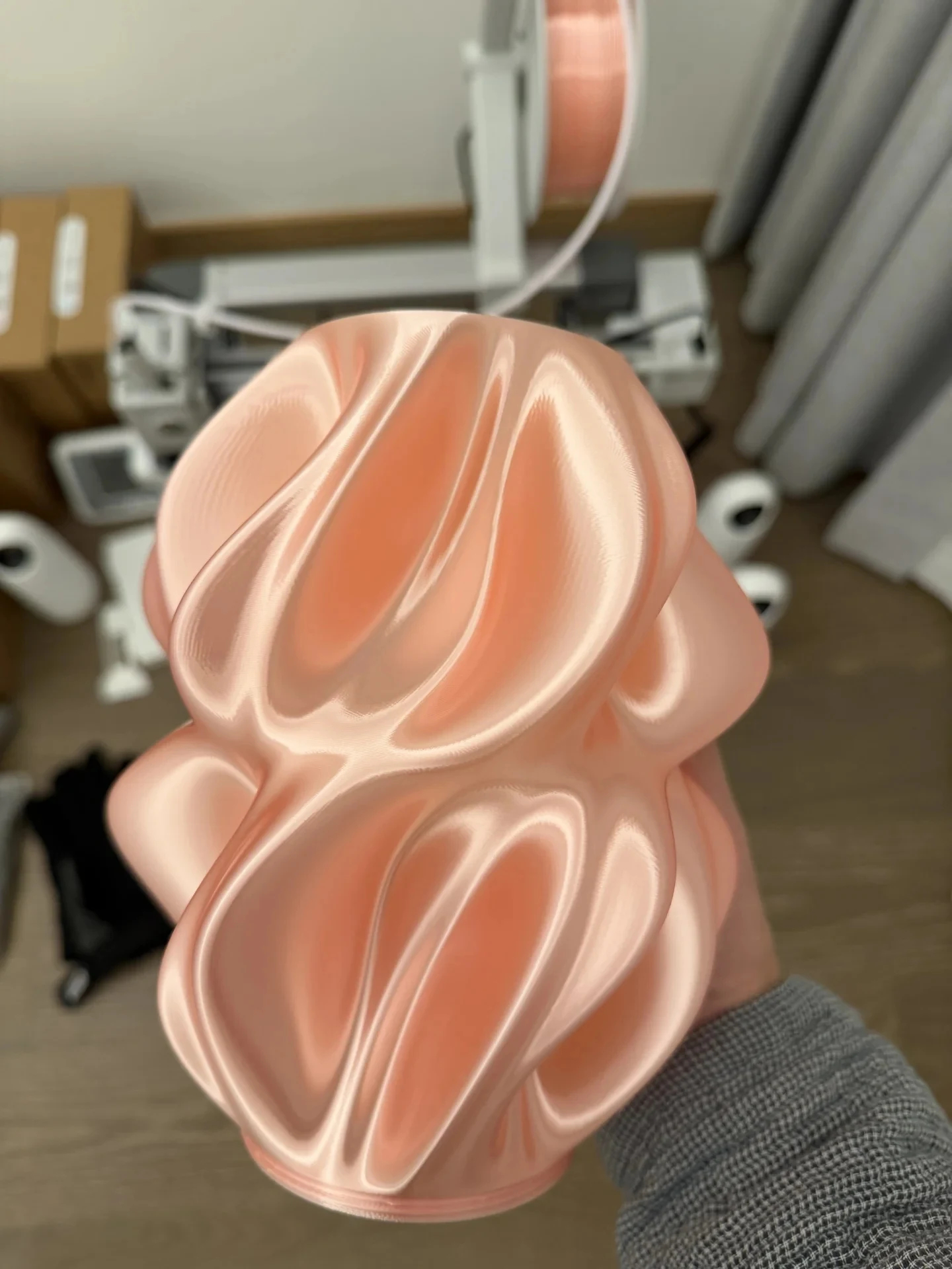 A hand holding a sculptural 3D-printed pink lamp shade with smooth, folded curves, positioned in front of an out-of-focus Bambu Lab A1 3D printer.