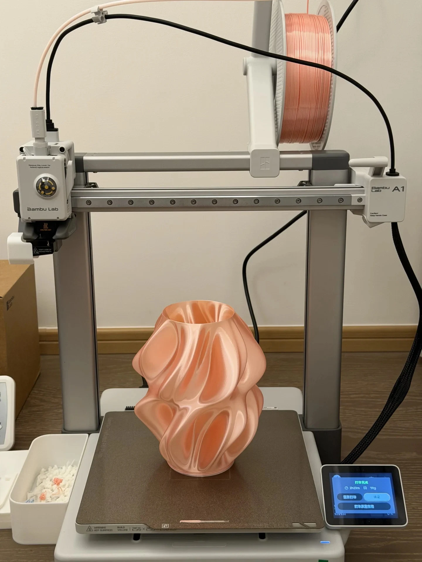Front view of a Bambu Lab A1 3D printer with a freshly printed silk pink twisted sculptural lamp body resting on the build plate beneath the toolhead, with an overhead spool of matching filament and an active touchscreen at the lower right.