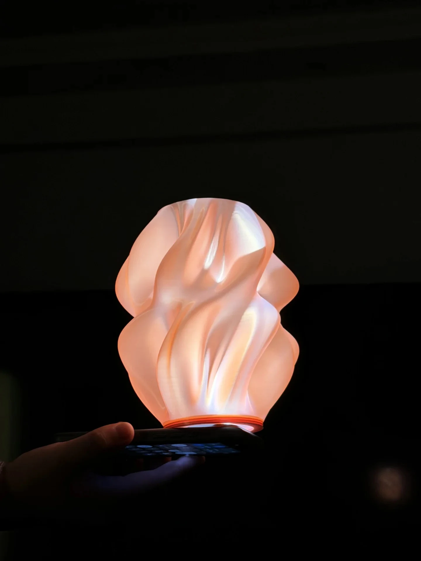 A 3D-printed lampshade with smooth, twisted organic ripples illuminated from underneath by a smartphone flashlight against a dark background.
