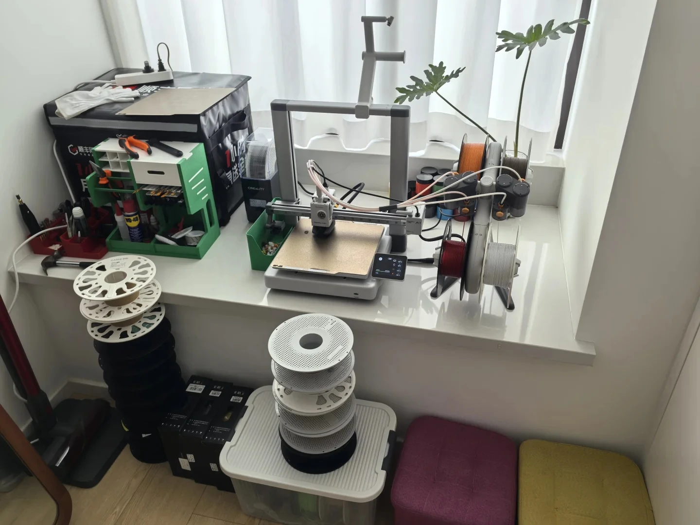 A Bambu Lab A1 3D printer sits on a wide white window ledge next to a four-spool feeder, tool caddies, and a filament dryer, with stacks of empty filament spools resting on the floor below.
