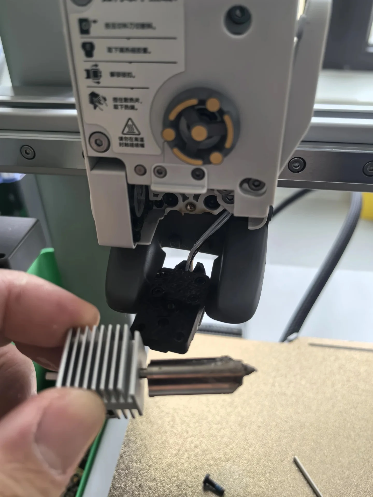 Close-up shot of the opened toolhead on a Bambu Lab A1 3D printer, with a hand holding the detached hotend and heatsink in the foreground and a removed screw lying on the build plate below.