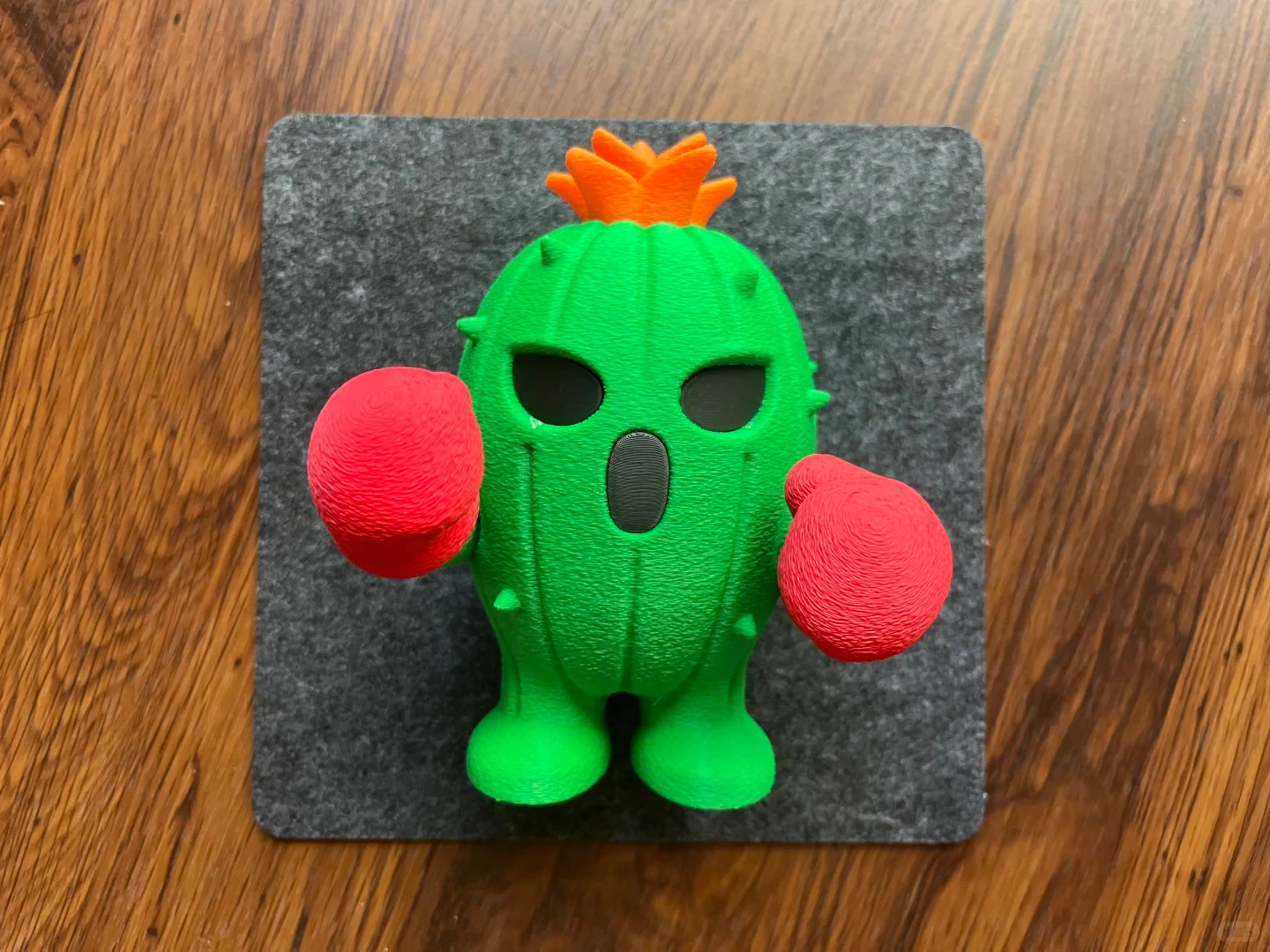 A 3D-printed green cactus figurine with red boxing gloves and an orange flower on its head, standing on a square gray felt pad set upon a wooden surface.