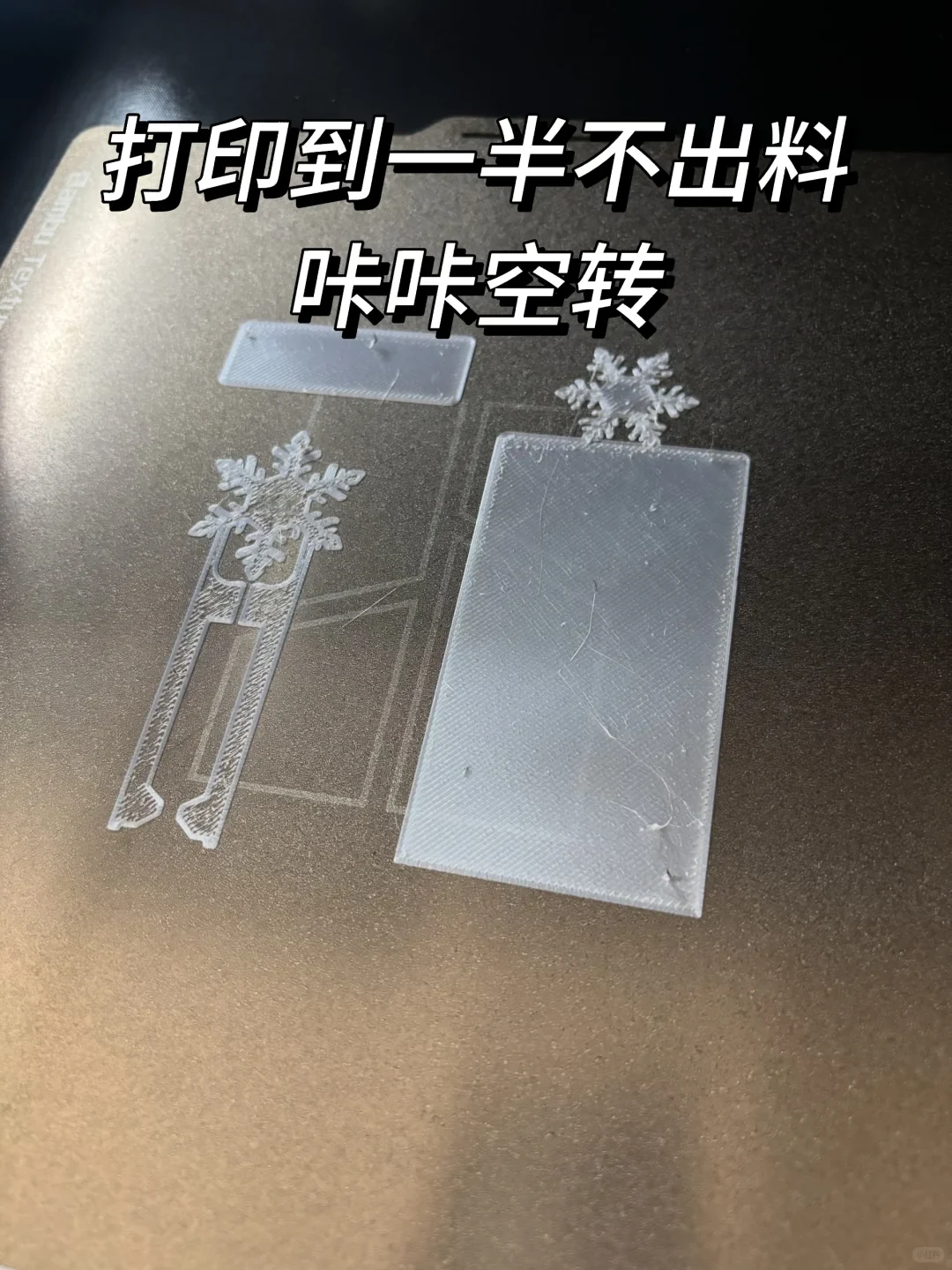 A 3D printer build plate showing an incomplete, failed print of snowflake-topped bookmarks in translucent white filament with text describing an extrusion failure.