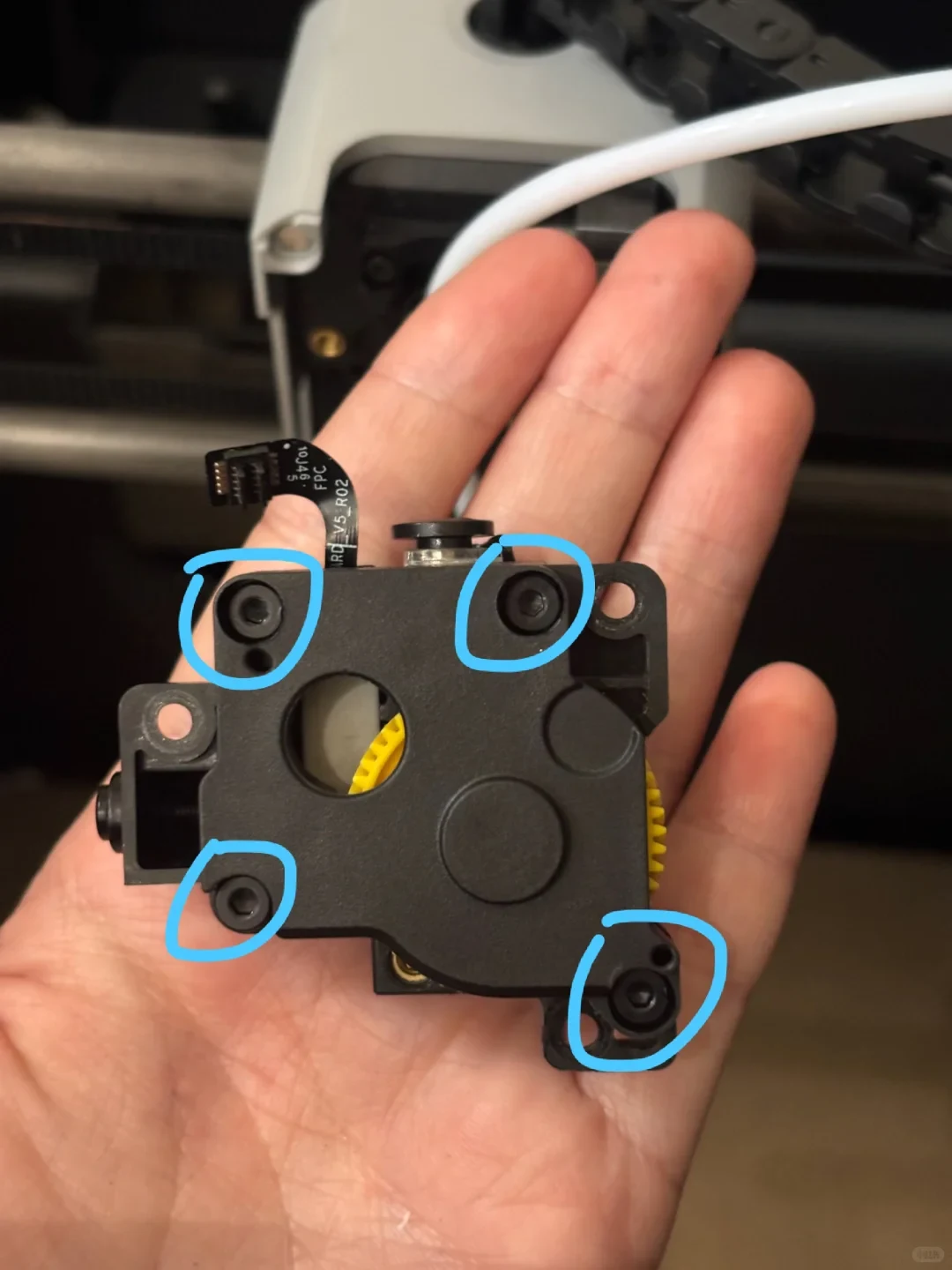 A person's hand holding a black 3D printer extruder mechanism, featuring four screw holes highlighted with light blue circles.