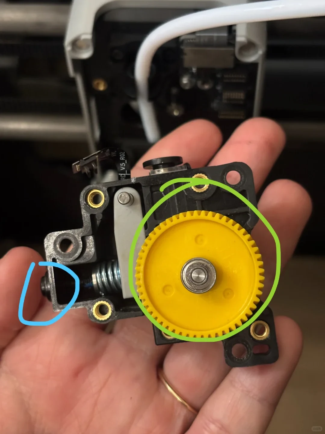A person holding a disassembled 3D printer extruder assembly, featuring a prominent yellow plastic gear, an internal tension spring, and threaded brass inserts, with a green circle drawn around the gear and a blue circle highlighting a side screw.