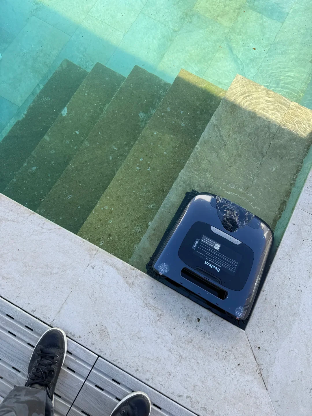 A Beatbot robotic pool cleaner resting on the shallow top step of a swimming pool beside the pool deck, with submerged steps covered in green algae visible beneath the water.