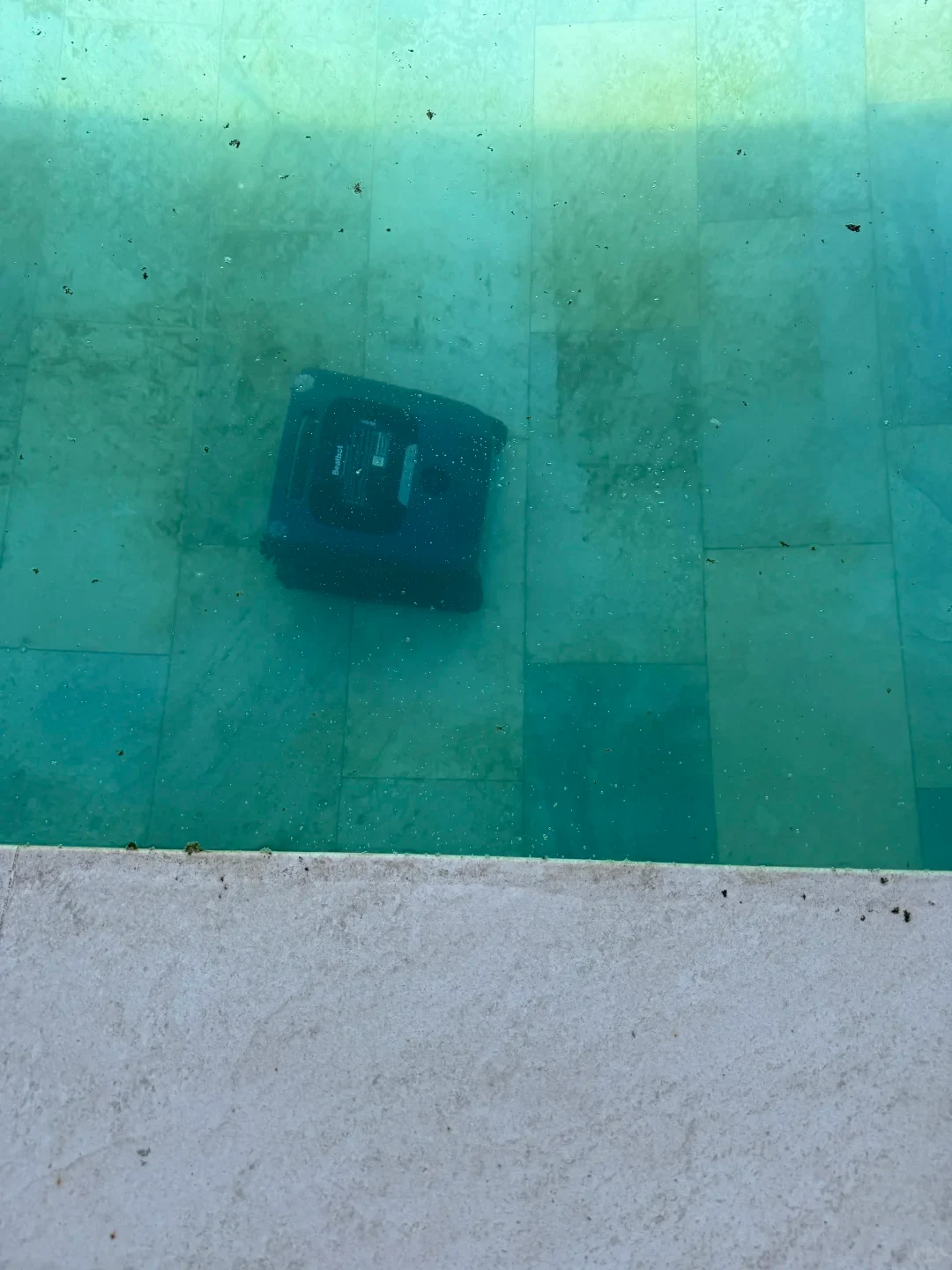 Top-down view of a dark Beatbot robotic pool cleaner submerged under greenish water on the tiled floor of a swimming pool, seen from the pool ledge.